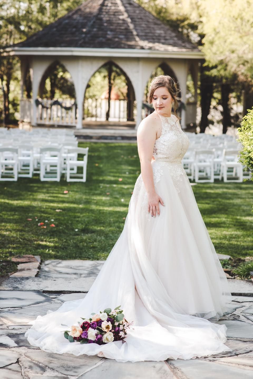 A wedding dress with lace details flows across a garden lawn with white chairs and a gazebo in the background.