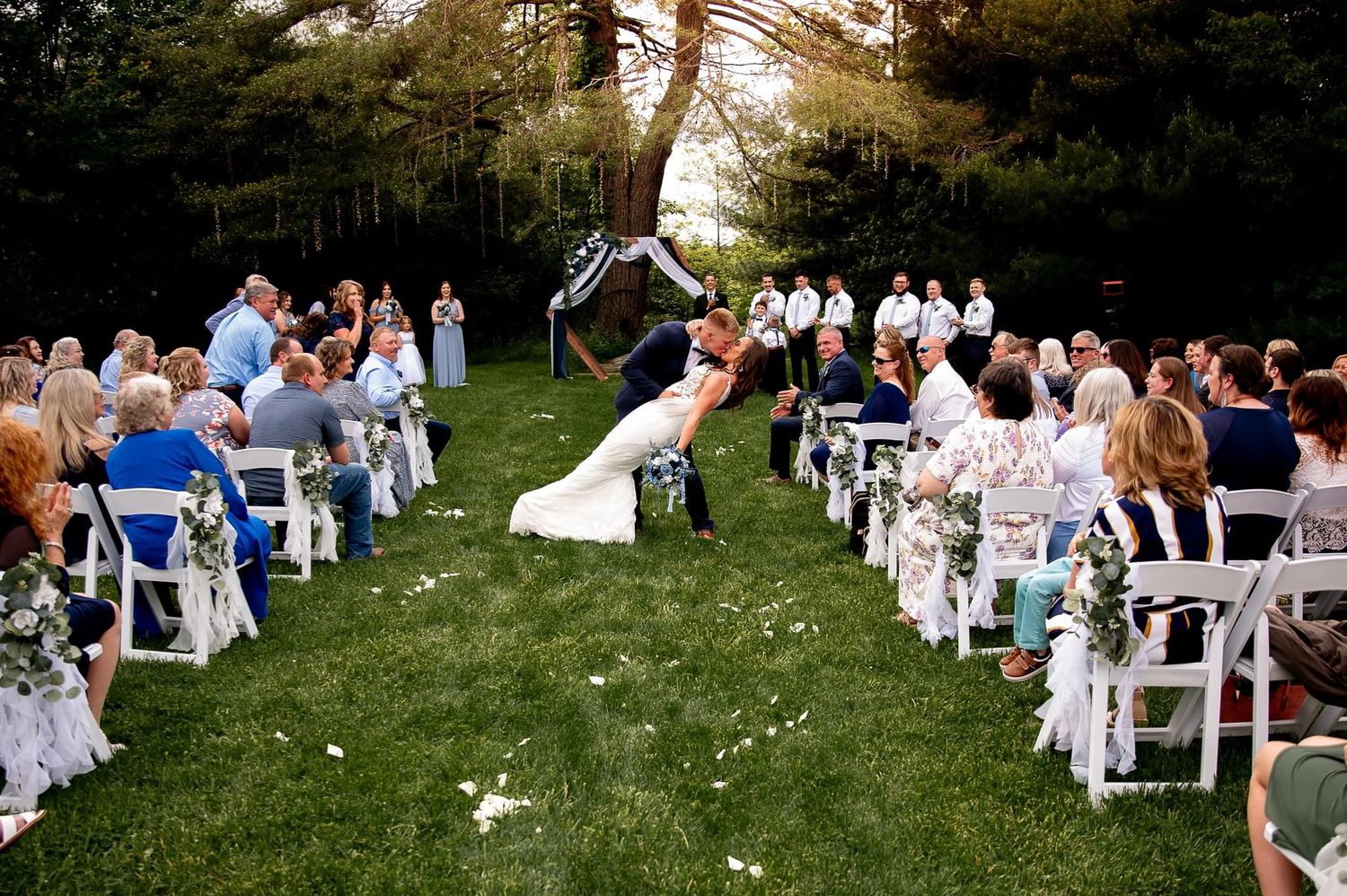 Wedding guests watch couple jumping in joy during outdoor ceremony with white chairs on grass.
