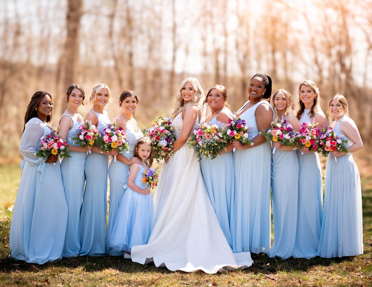 Wedding party in light blue dresses holding vibrant bouquets outdoors.