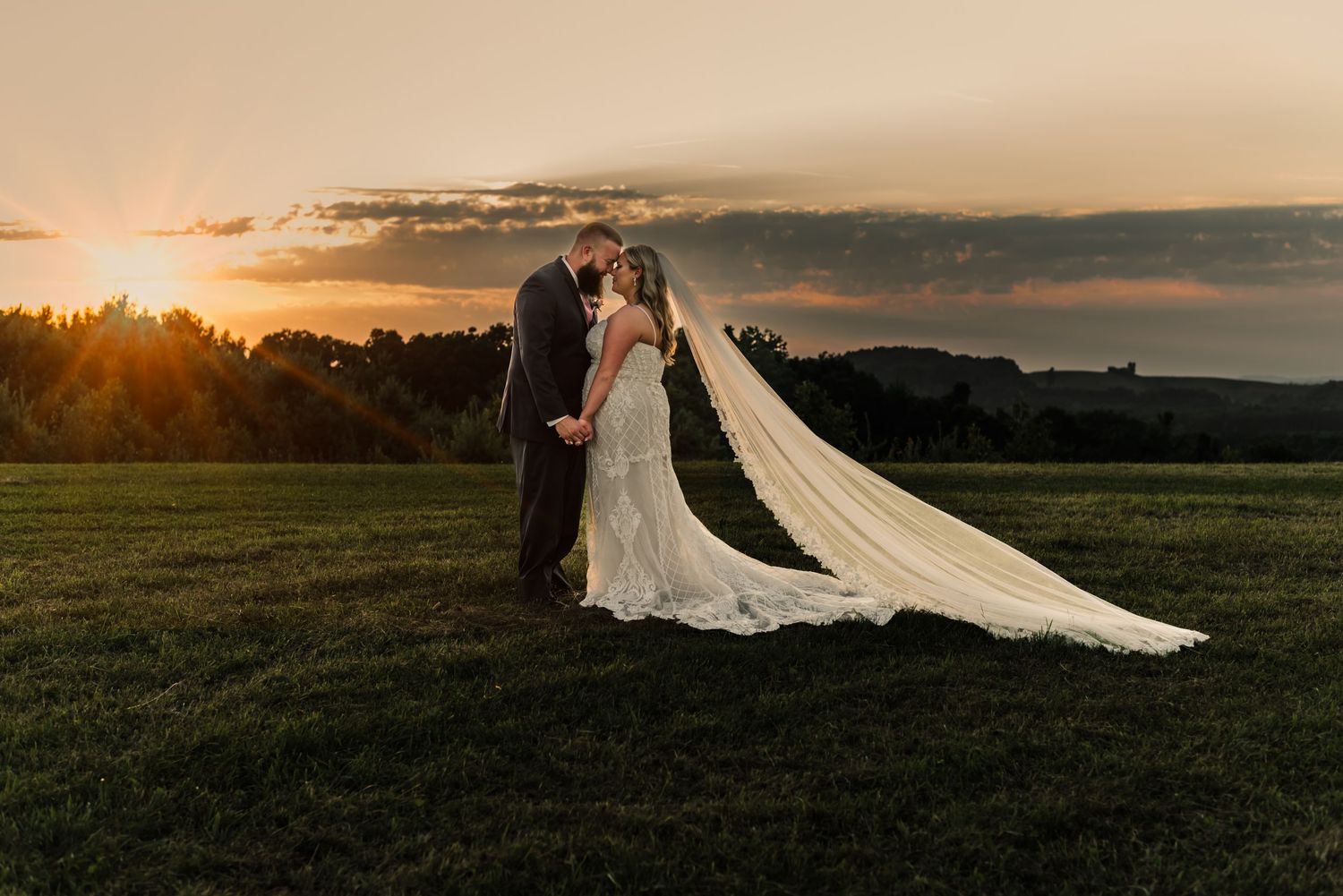 A dramatic sunset wedding photo with a flowing cathedral veil against an orange sky.