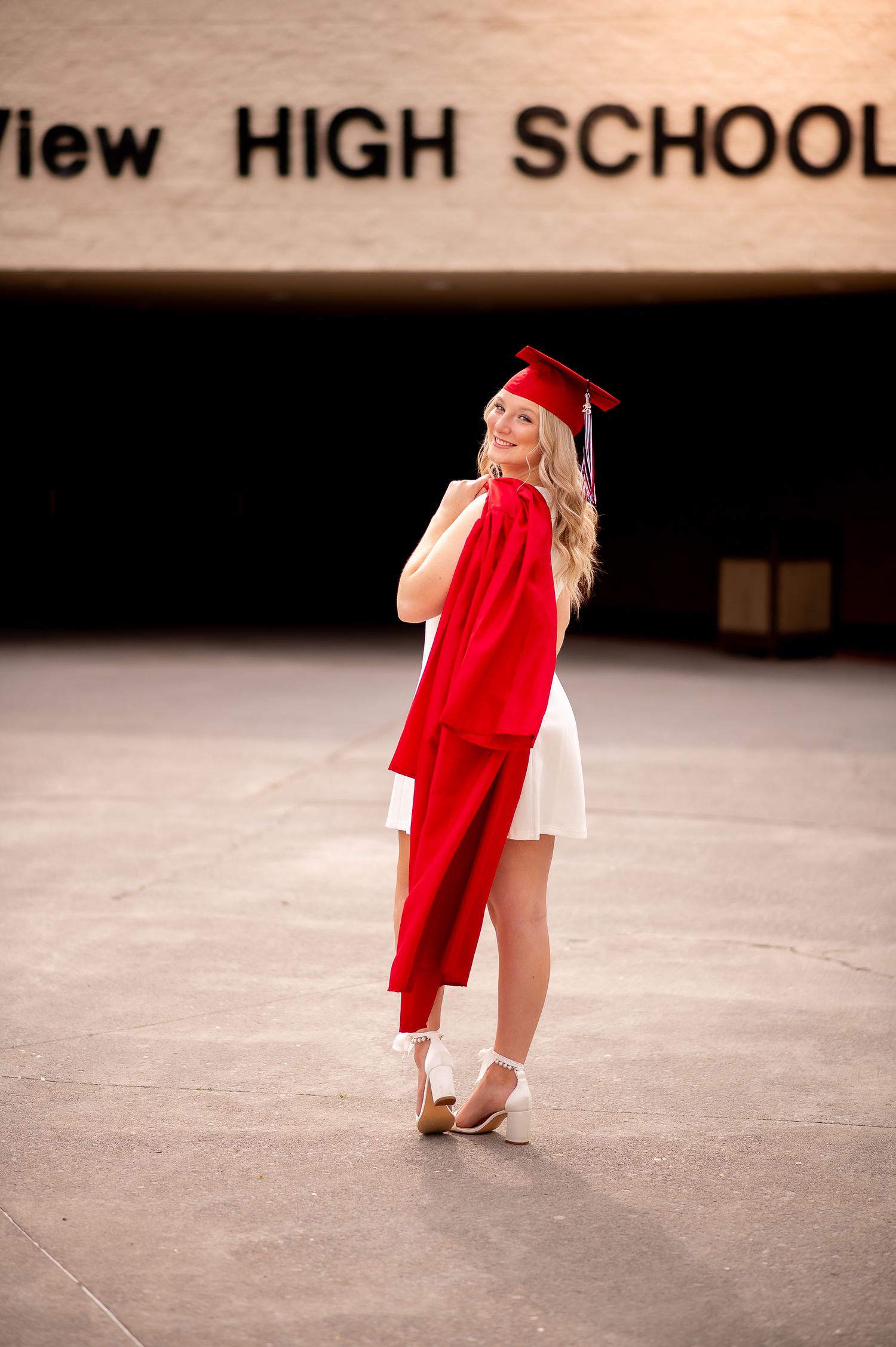 A graduate in red cap and gown poses in front of their high school building.