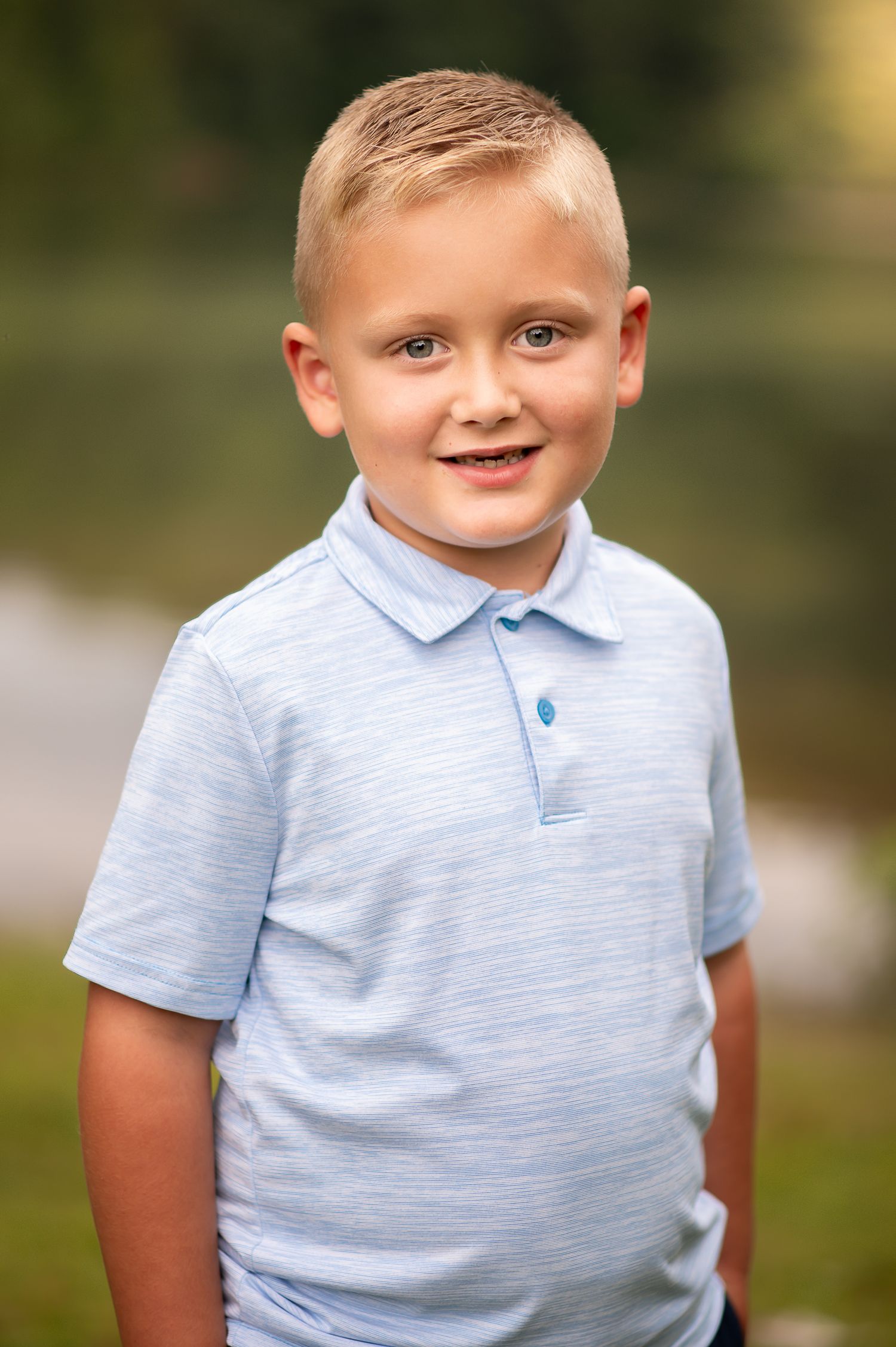 Young child wearing a light blue polo shirt smiling during an outdoor portrait session.