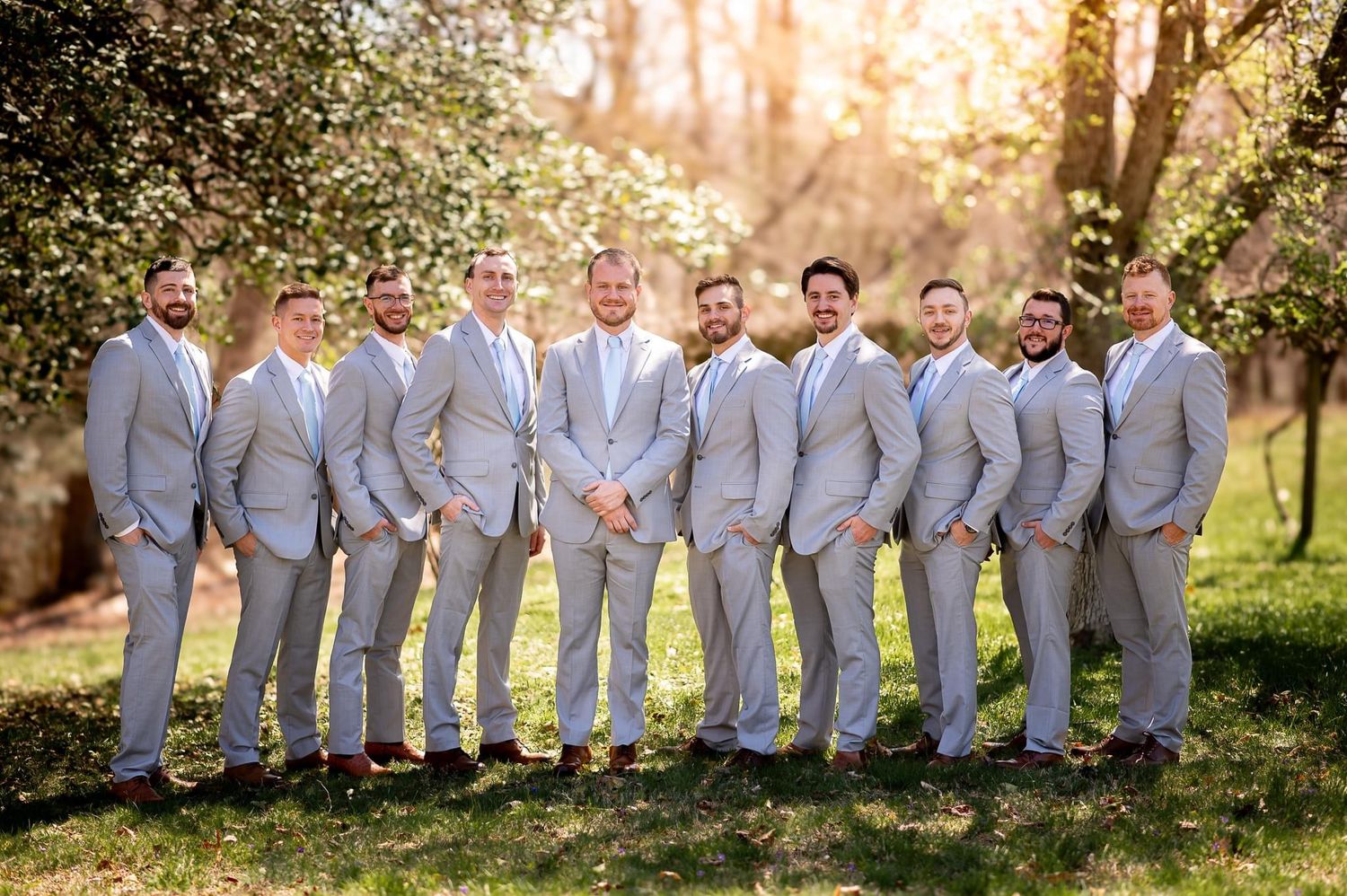 Groomsmen in matching light gray suits pose outdoors in natural lighting.
