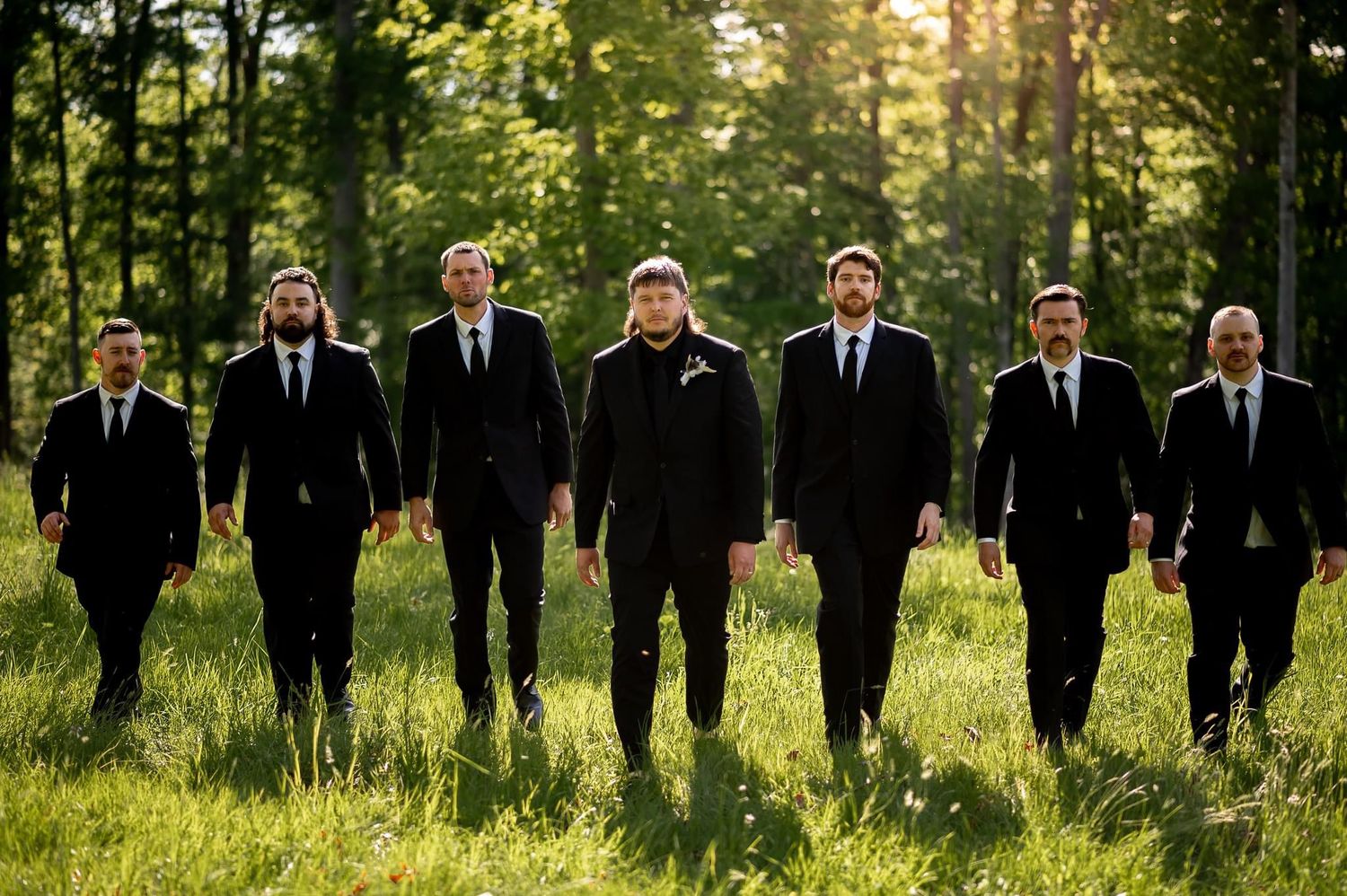 Wedding party members in dark suits walk through sunlit grassy field.