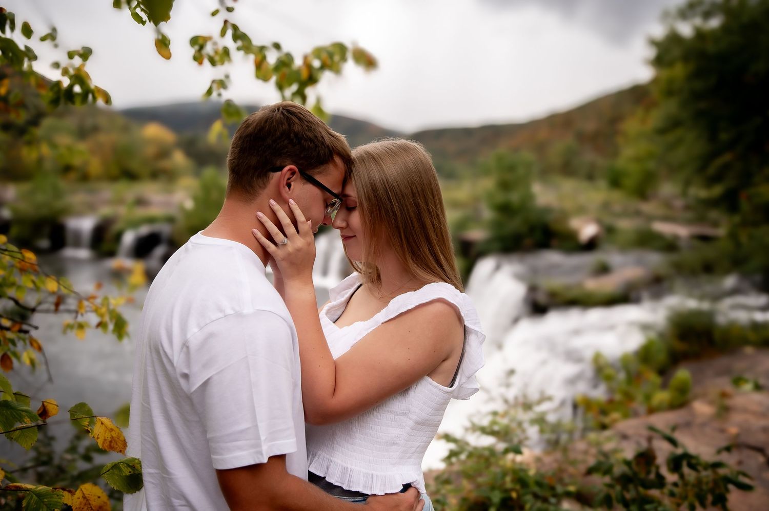 Romantic couple embracing near a flowing river with mountains in the background.