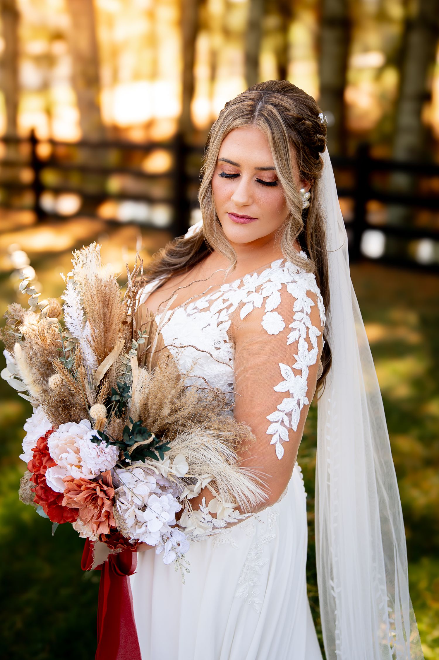 Bride in lace wedding dress holds romantic bouquet with pampas grass and white flowers in golden sunlit forest.
