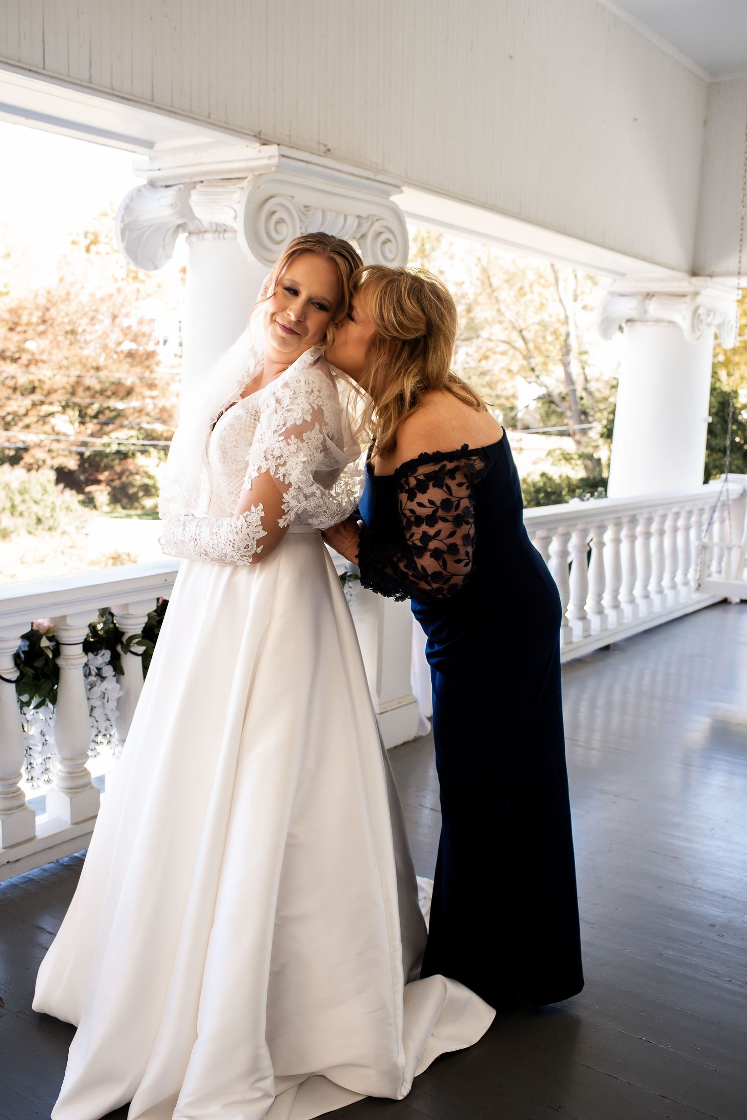 Two people share an emotional moment on a white columned porch during a wedding.