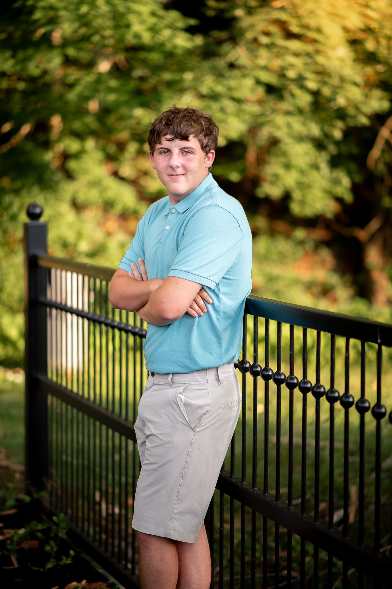 Casual outdoor portrait by metal fence wearing light blue polo and khaki shorts.