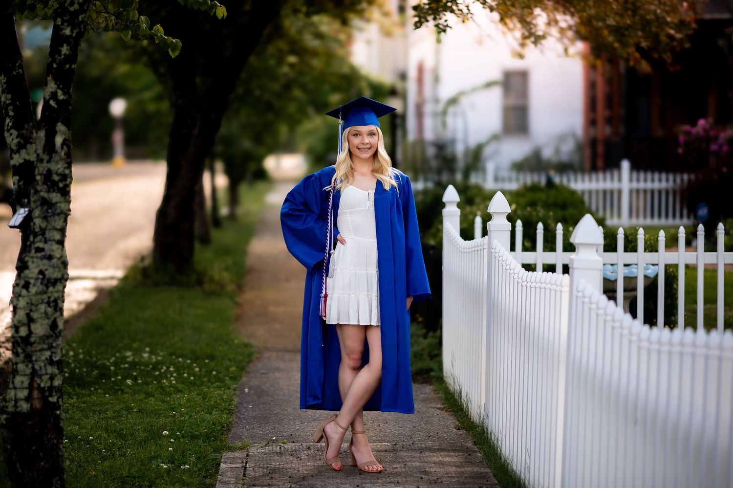 A graduation portrait in blue cap and gown standing by a white picket fence at sunset.