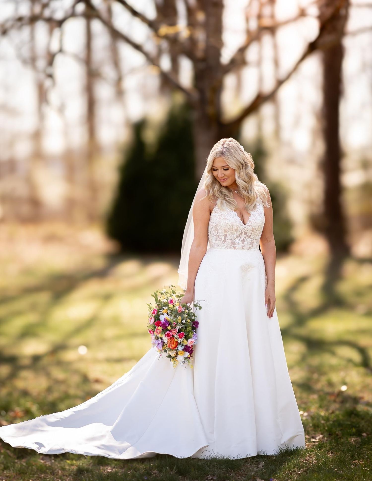 A bride in a flowing white gown holds a colorful bouquet in a natural outdoor setting.