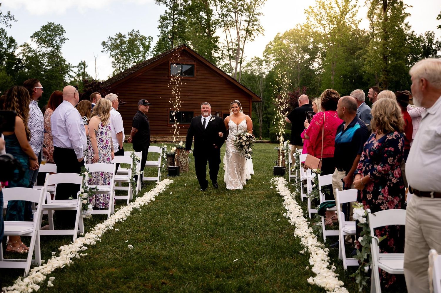 Outdoor wedding ceremony at rustic venue with white chairs and wooden lodge in background.