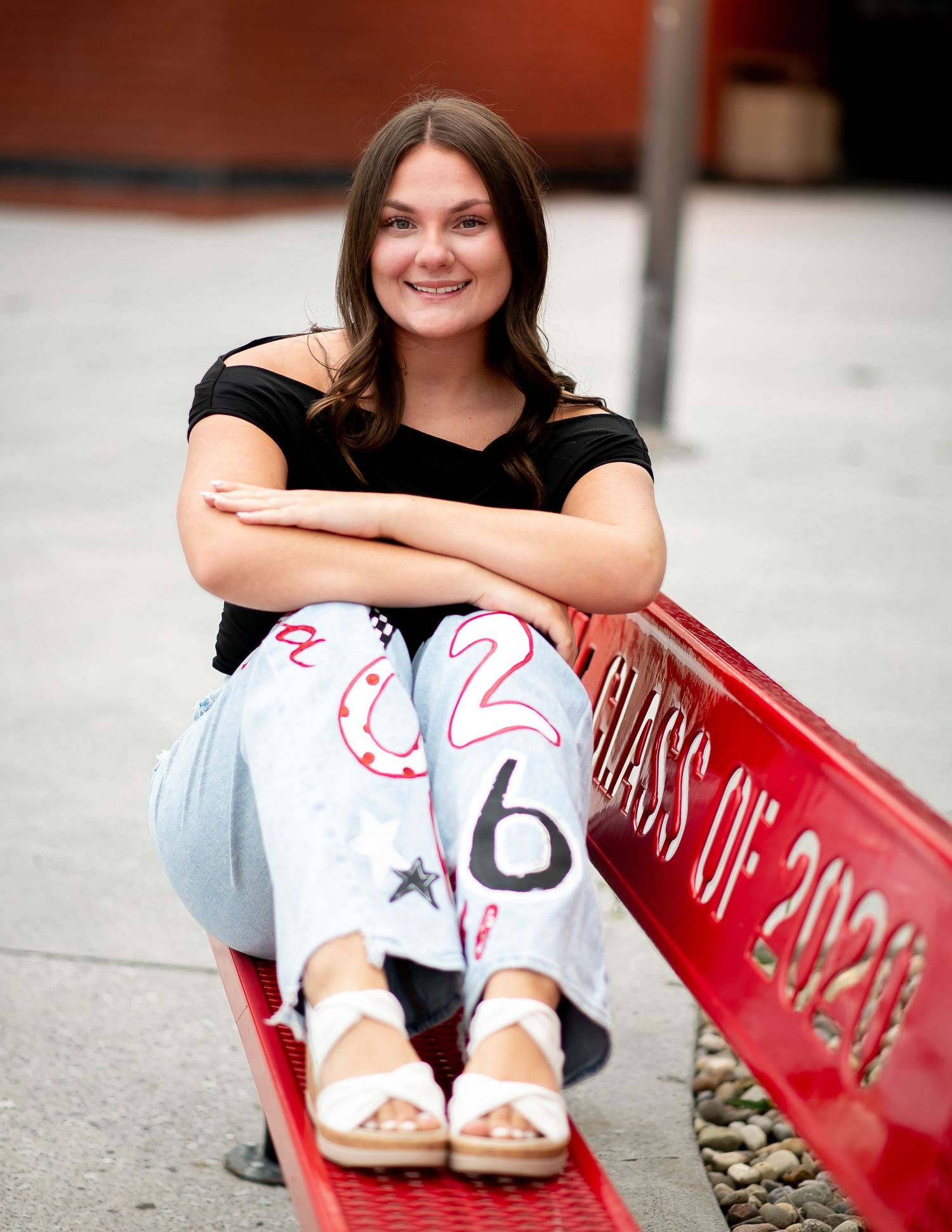 High school senior portrait features a casual pose on a red bench wearing light jeans and a black top with sandals.