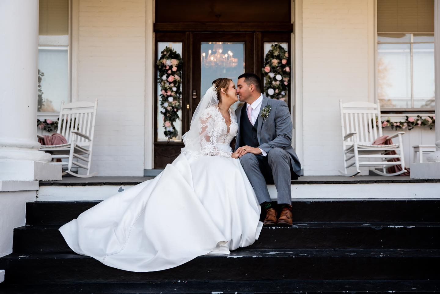 A newlywed couple poses together on a grand white porch with pillars.