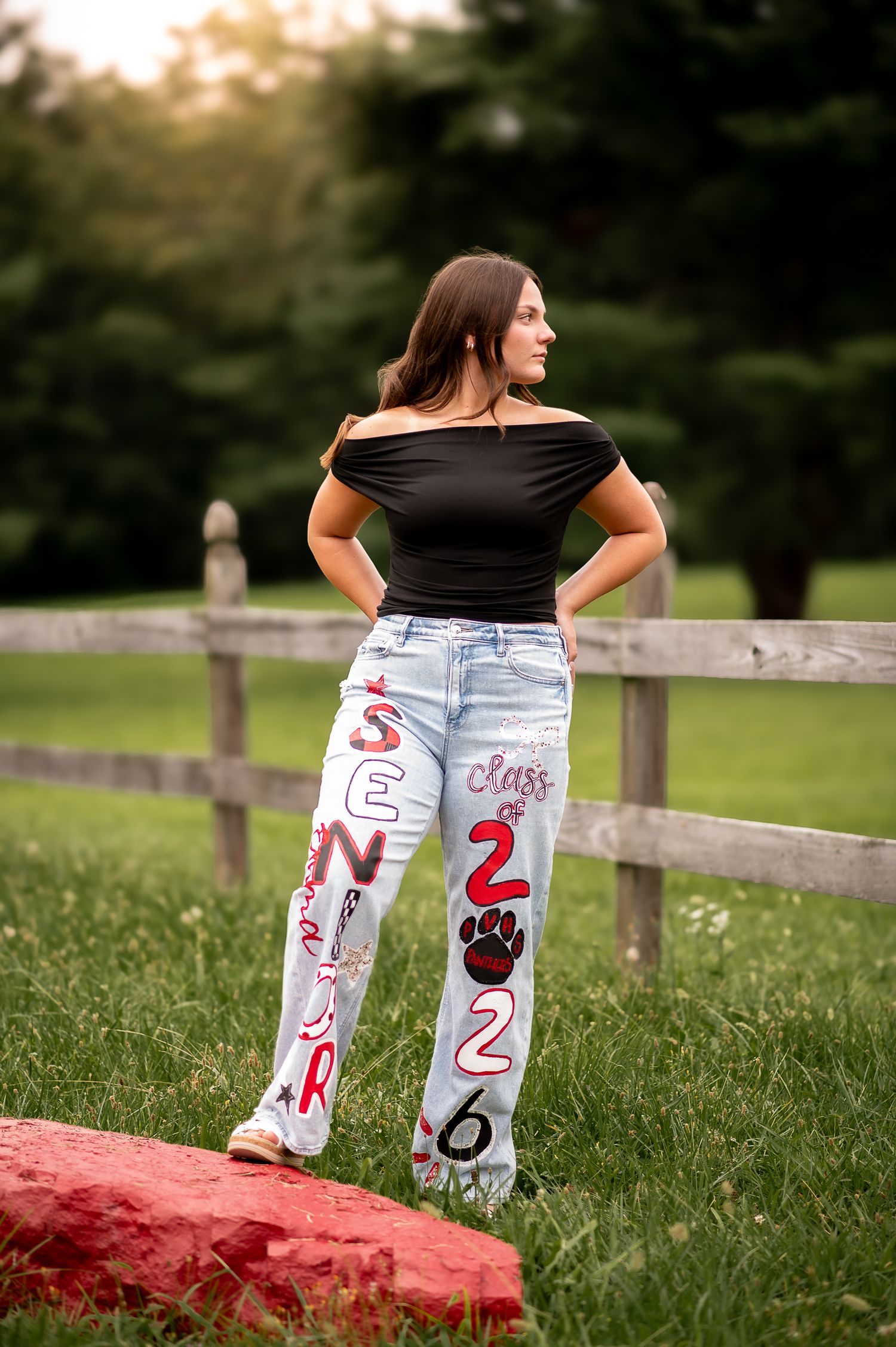 Standing against a rustic farm fence at sunset, someone poses in a black off-shoulder top and decorated light denim jeans.