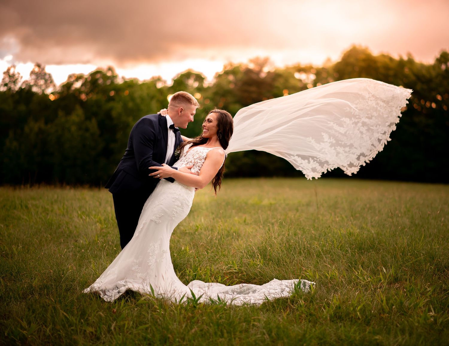 A romantic wedding photo in a field at sunset with the bride's veil dramatically flowing in the wind.