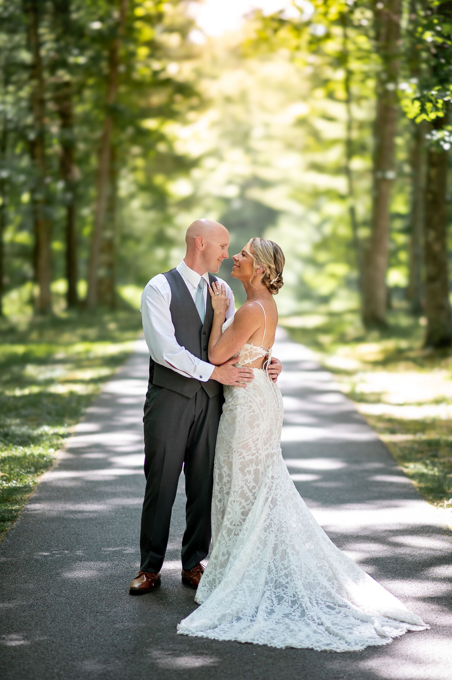 A romantic wedding photo of a couple embracing on a sunlit forest path with a flowing lace dress train.