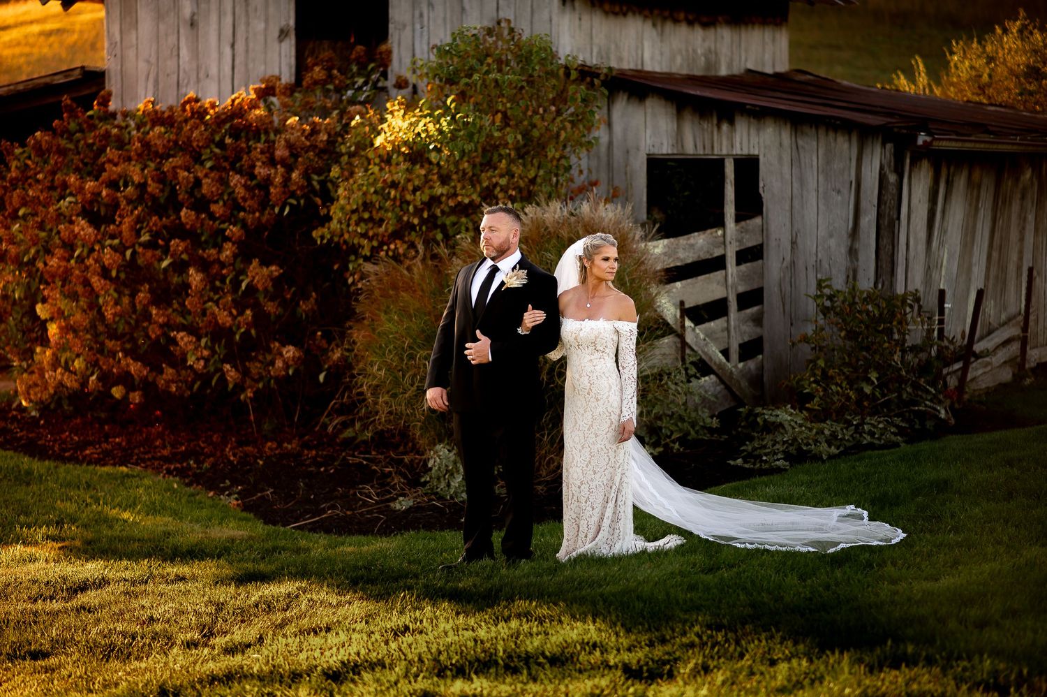 Couple in wedding attire walking past a rustic barn during sunset.