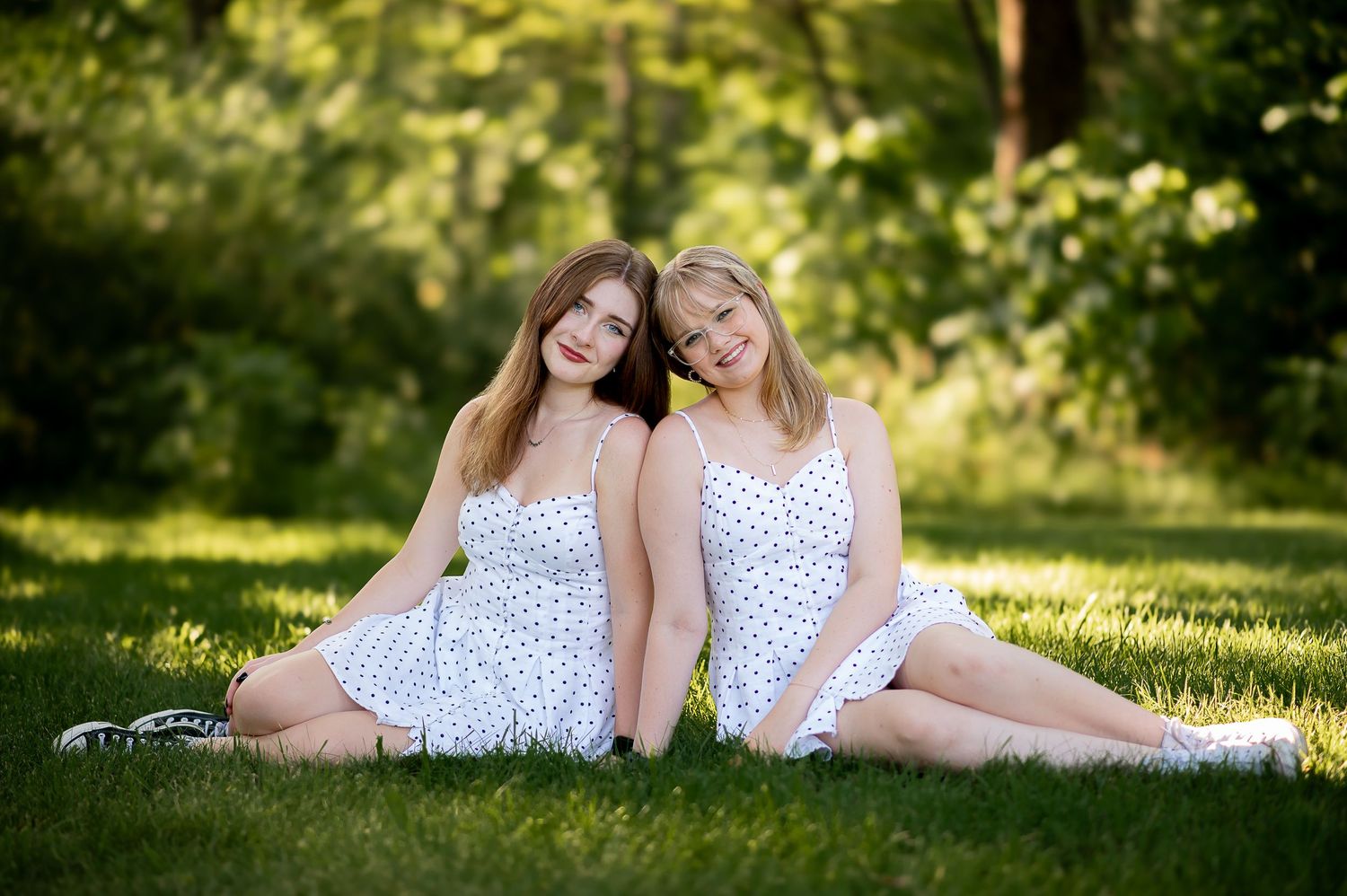 Two people in matching polka dot dresses pose together on grass in a sunny garden setting.