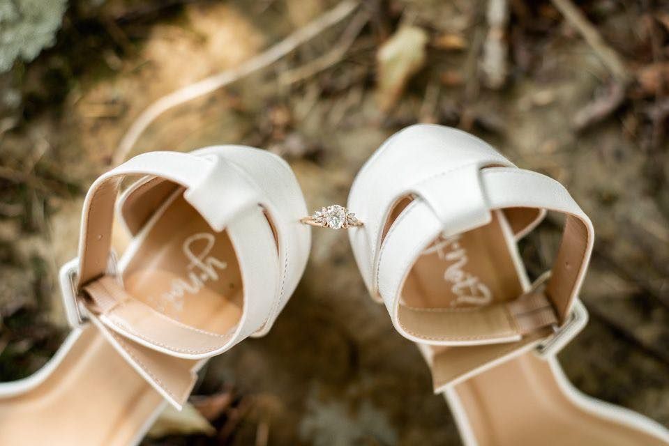 White wedding heels display an engagement ring between them on natural ground.
