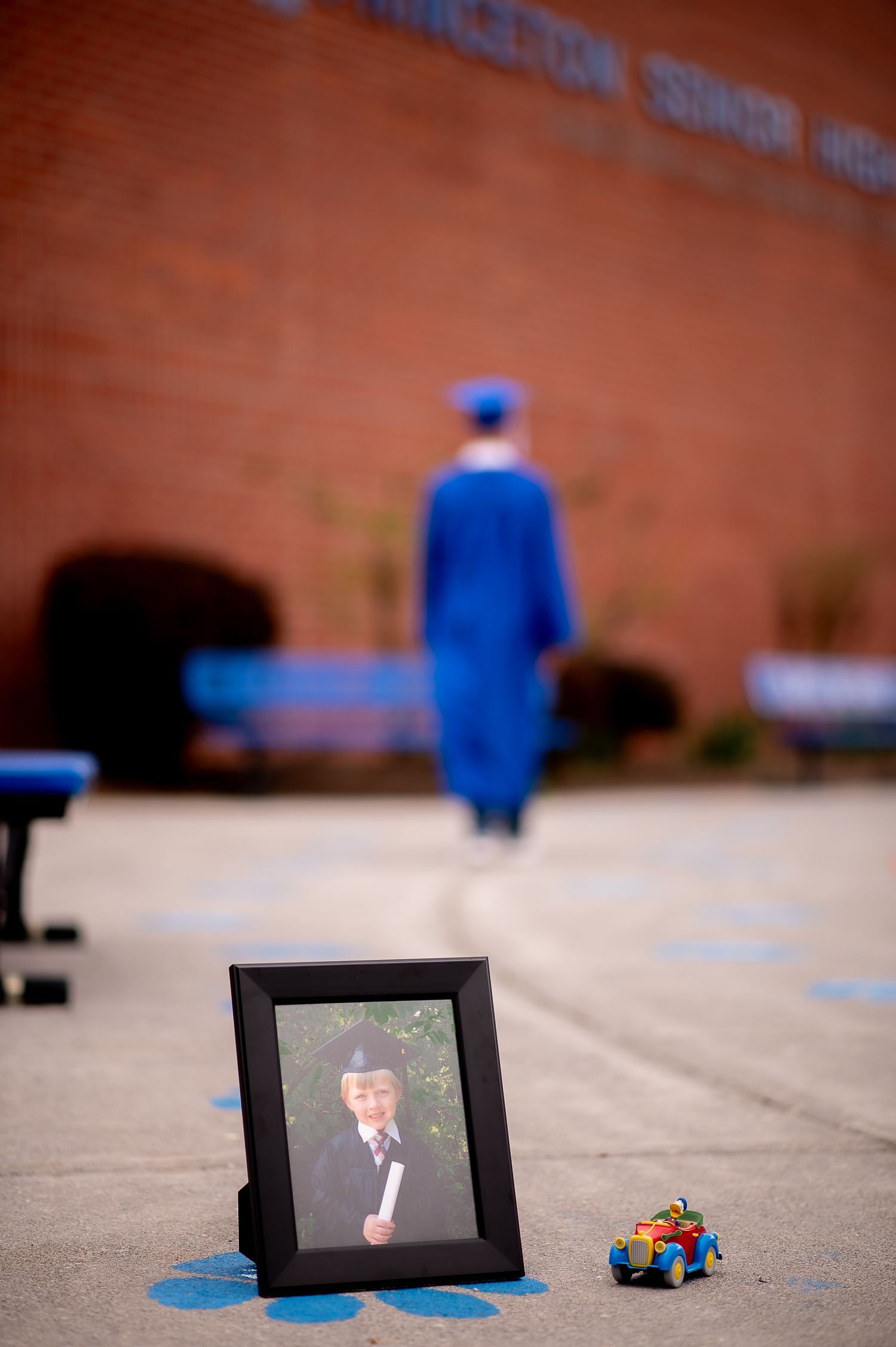 A graduation photo setup with a framed portrait and blue toy car in front of a blurred graduate.