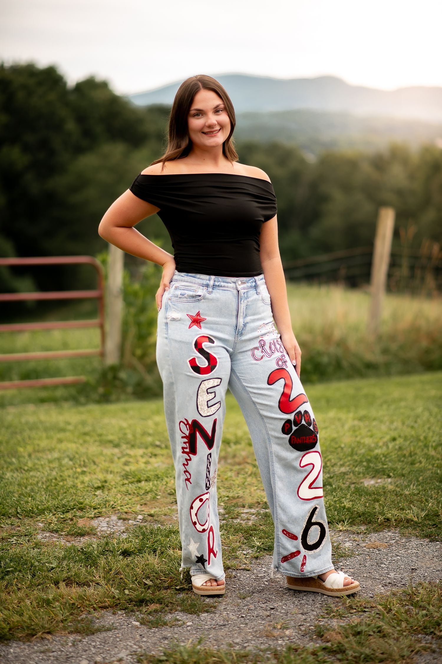 Off-shoulder black top paired with customized light denim jeans featuring hand-painted designs against a scenic mountain backdrop.