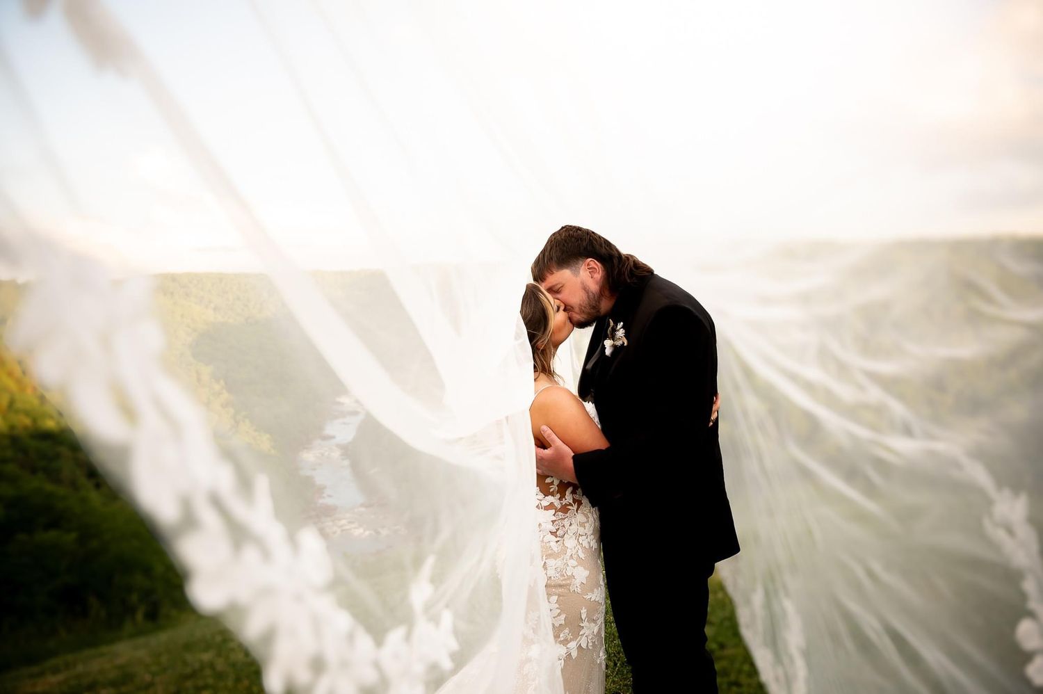 Romantic wedding portrait with flowing veil against sunset sky.