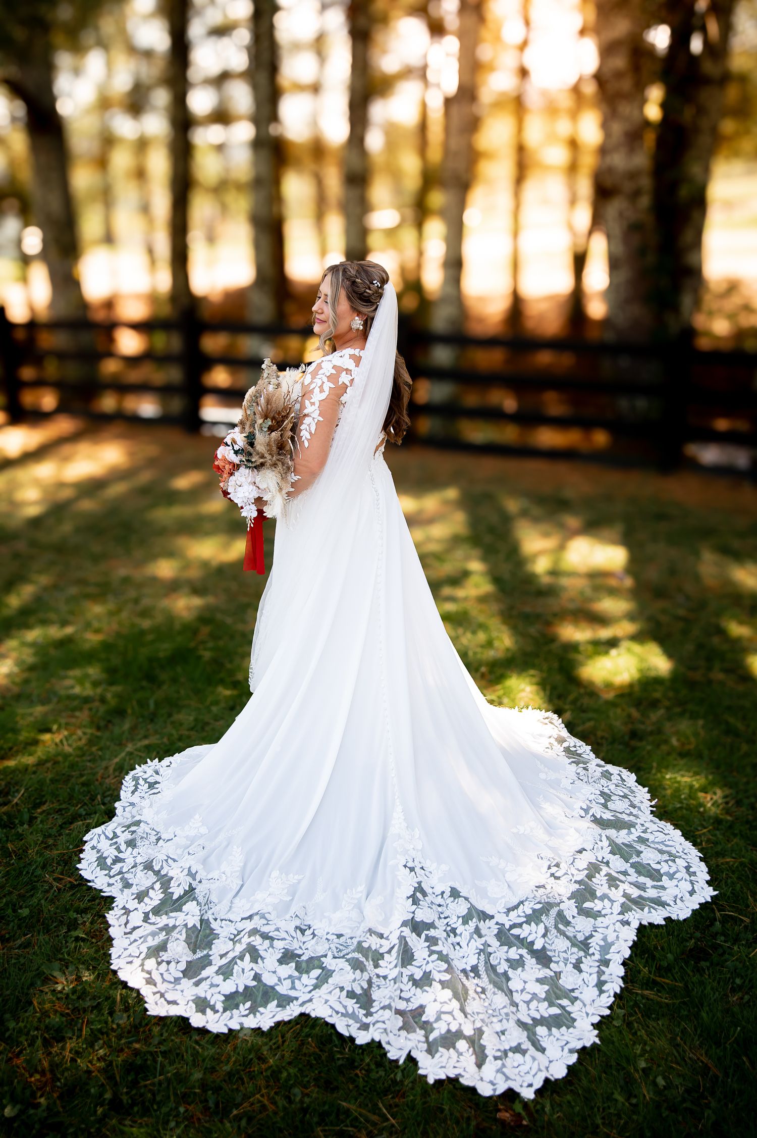 A flowing white wedding dress with intricate lace train spreads elegantly across sunlit grass during golden hour.