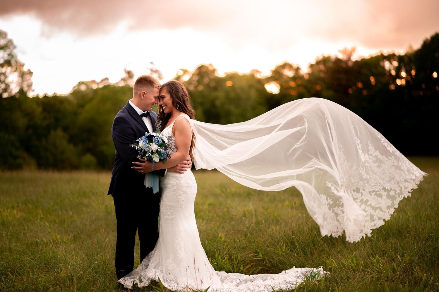 Wedding couple embraces in a field at sunset with flowing lace veil creating romantic silhouette.