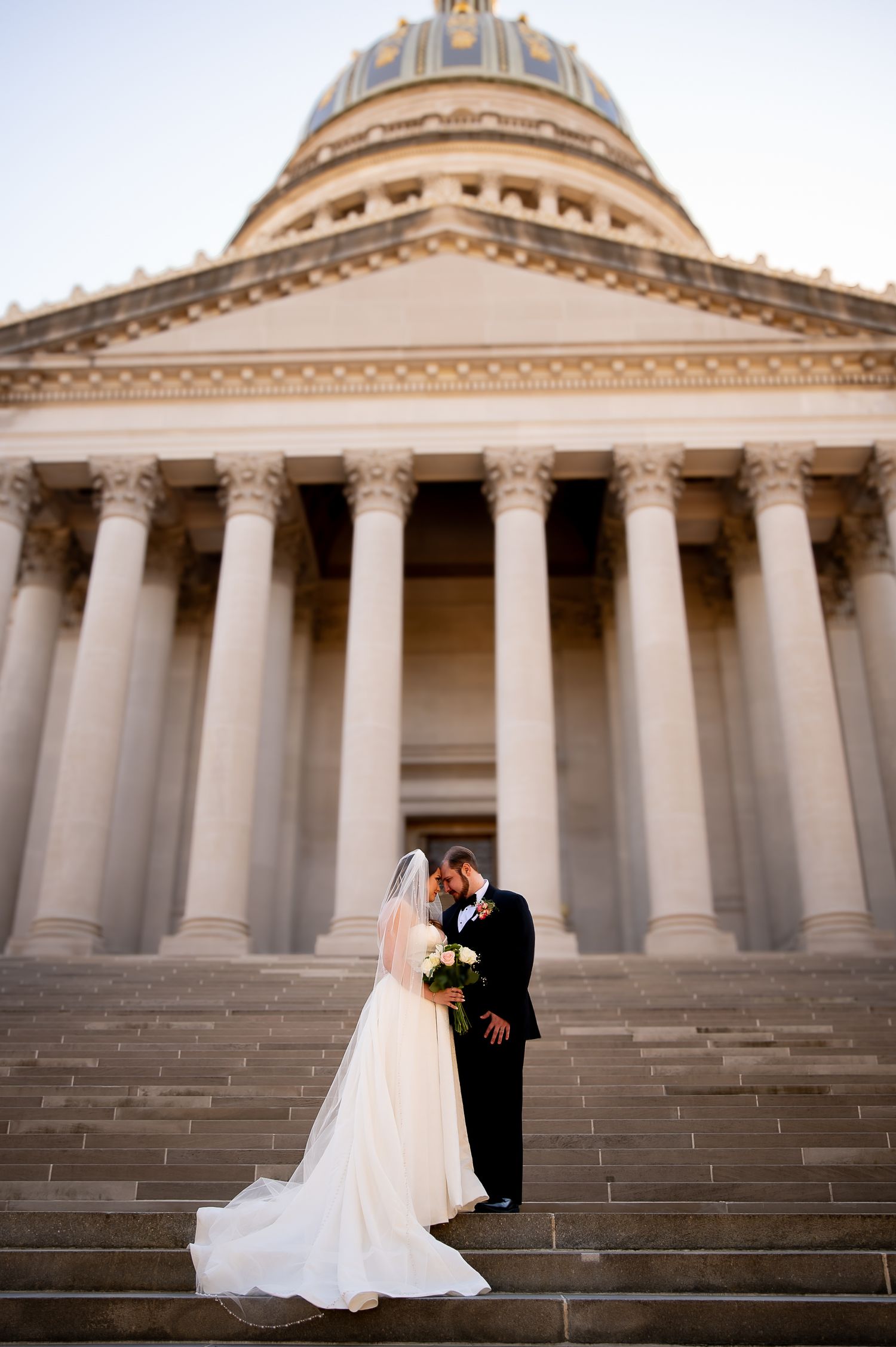 Wedding couple poses on grand stone steps of a neoclassical domed building.