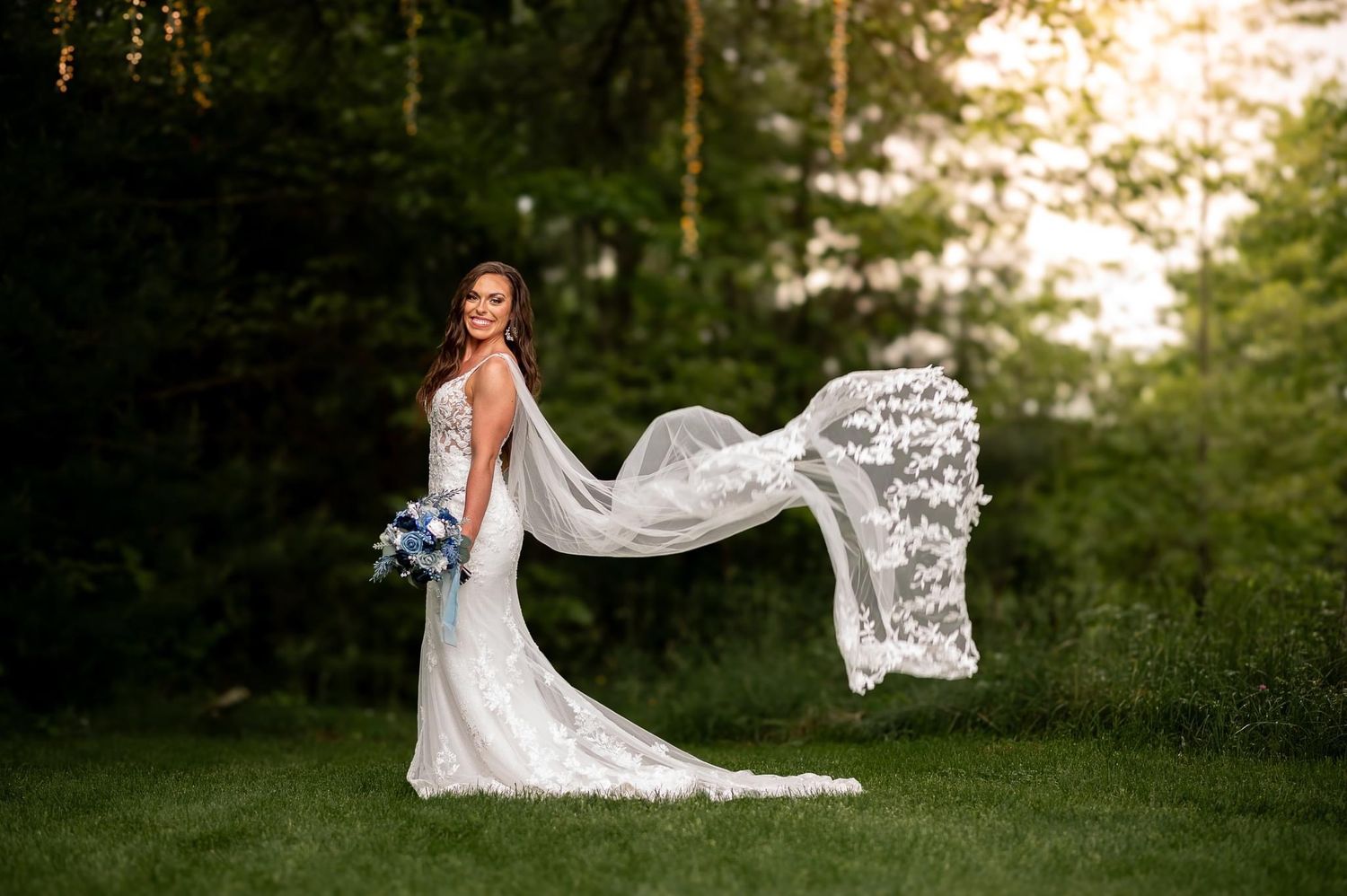 Elegant bride in lace gown with flowing veil poses in garden setting at sunset.