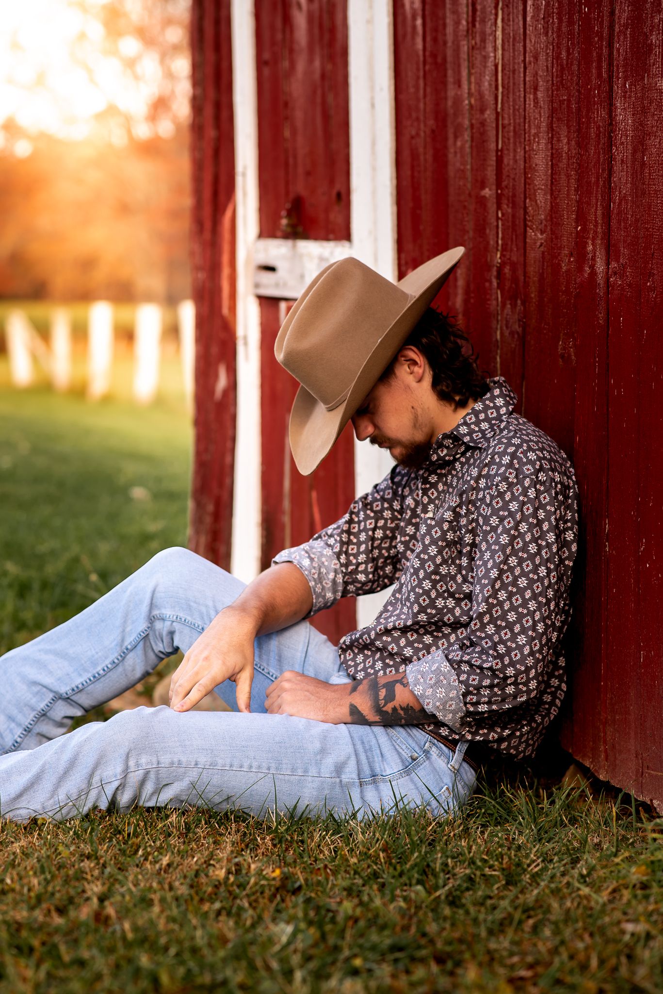 Cowboy in denim and patterned shirt relaxing against red barn wall at sunset.