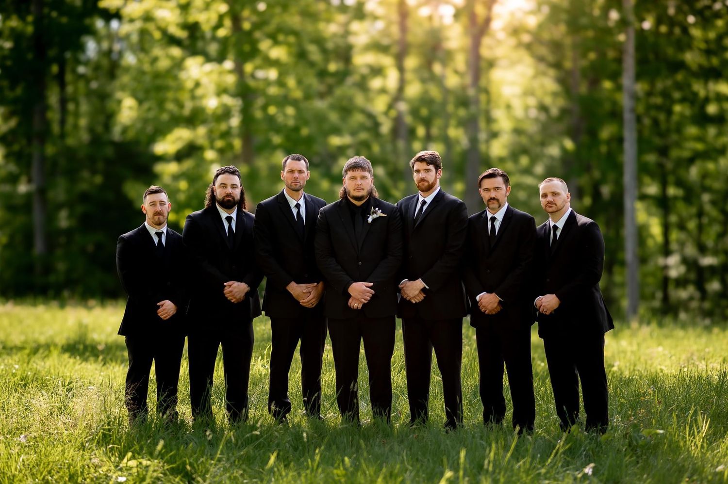 Groomsmen in black suits pose together on sunny grass field.