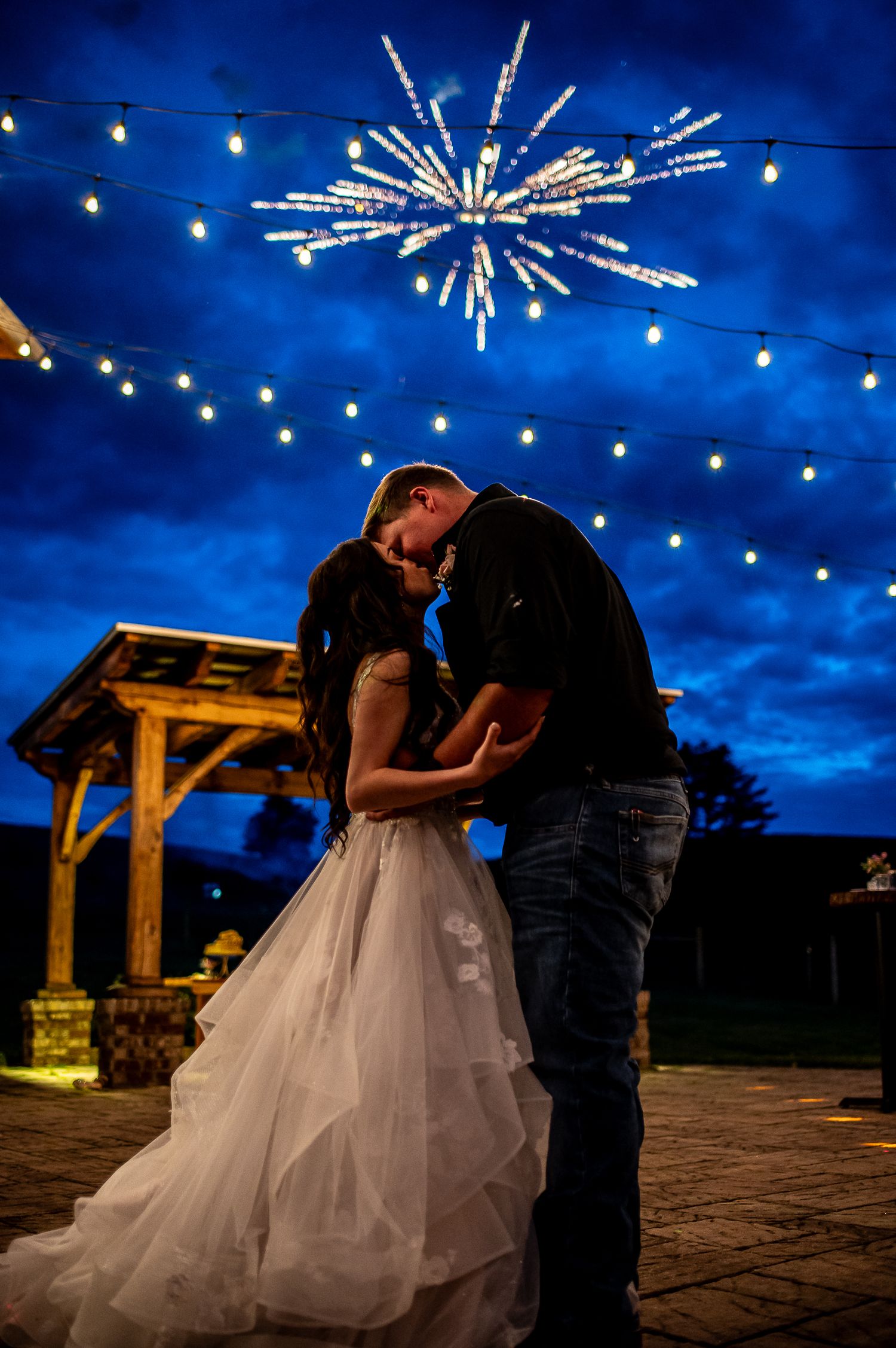 A romantic kiss under twinkling lights and fireworks at dusk.