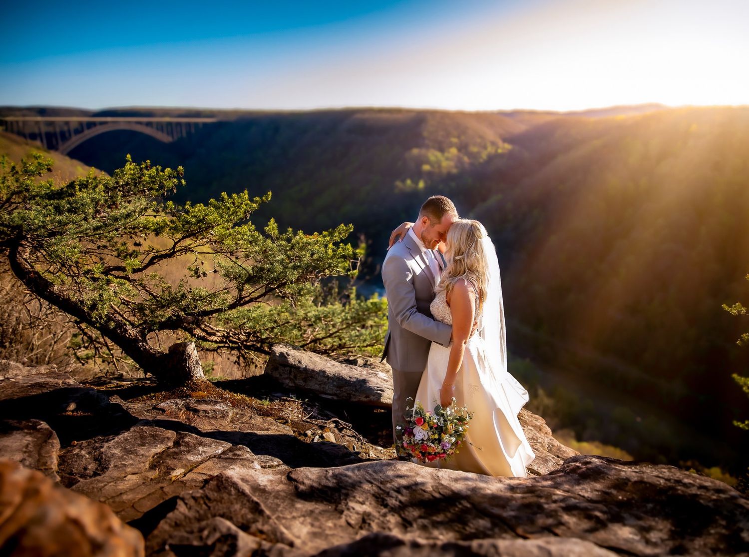 Wedding couple poses on scenic mountain overlook against bright blue sky and sunburst.