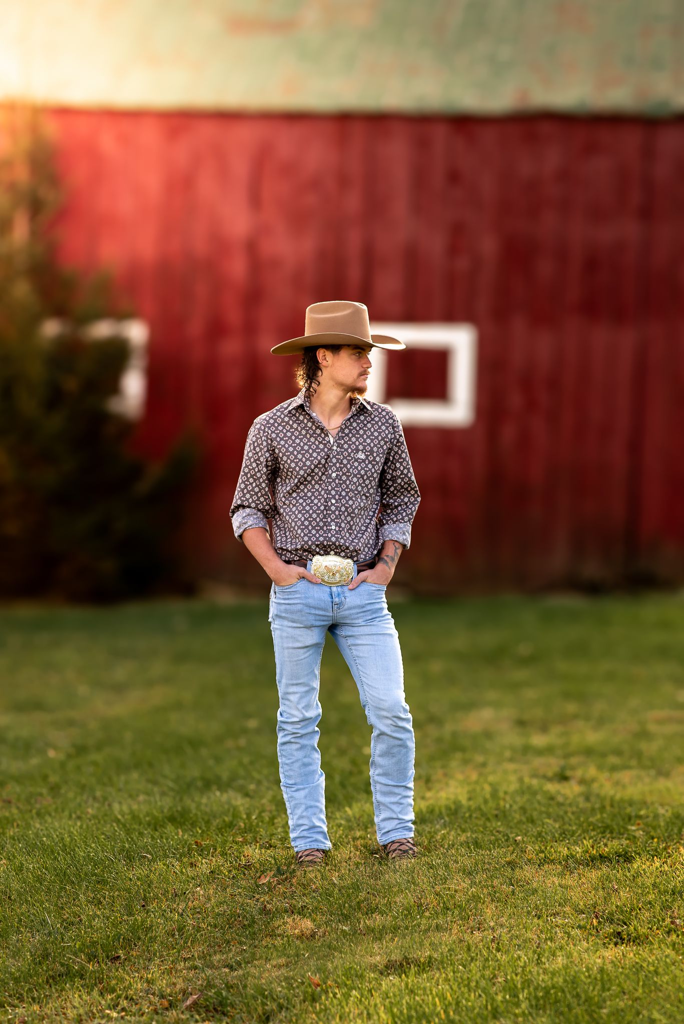 Western style portrait of person in cowboy hat and patterned shirt standing by red barn.