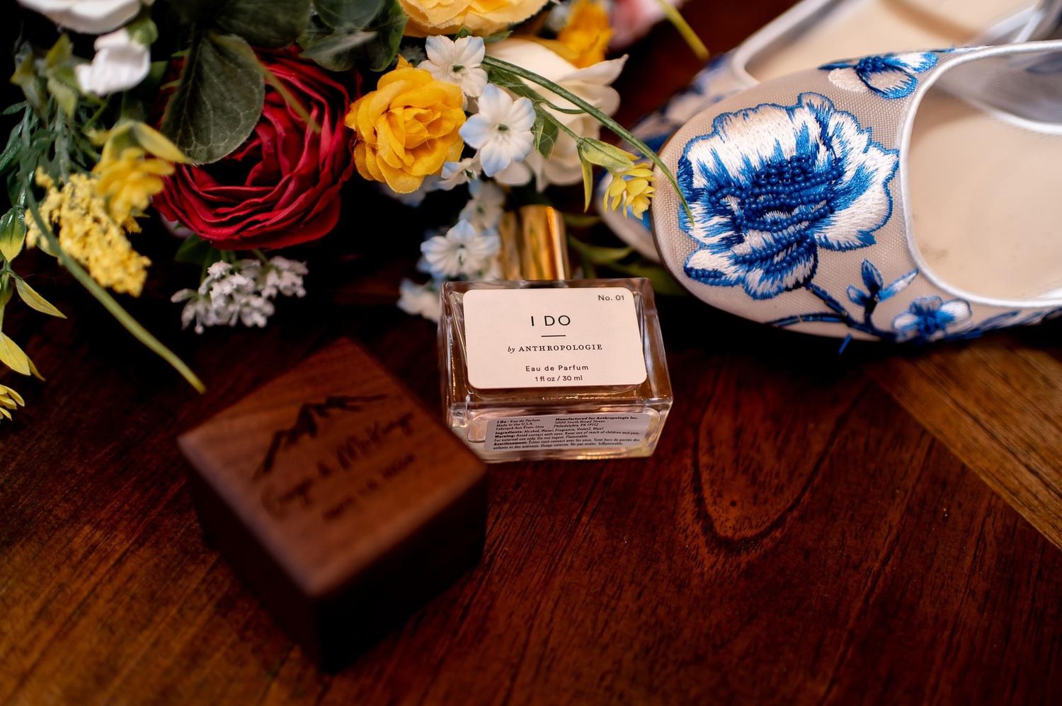Wedding details featuring perfume bottle, blue floral shoe, and roses on wooden surface.