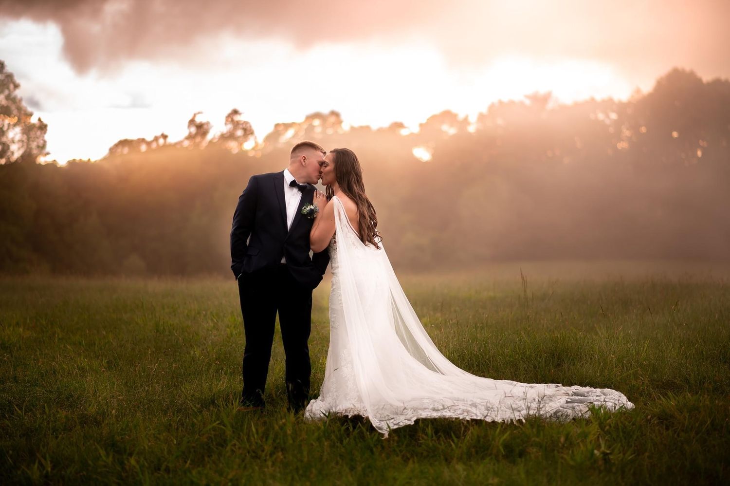 A romantic wedding photo in a misty field at sunset with a long flowing white dress trailing across the grass.