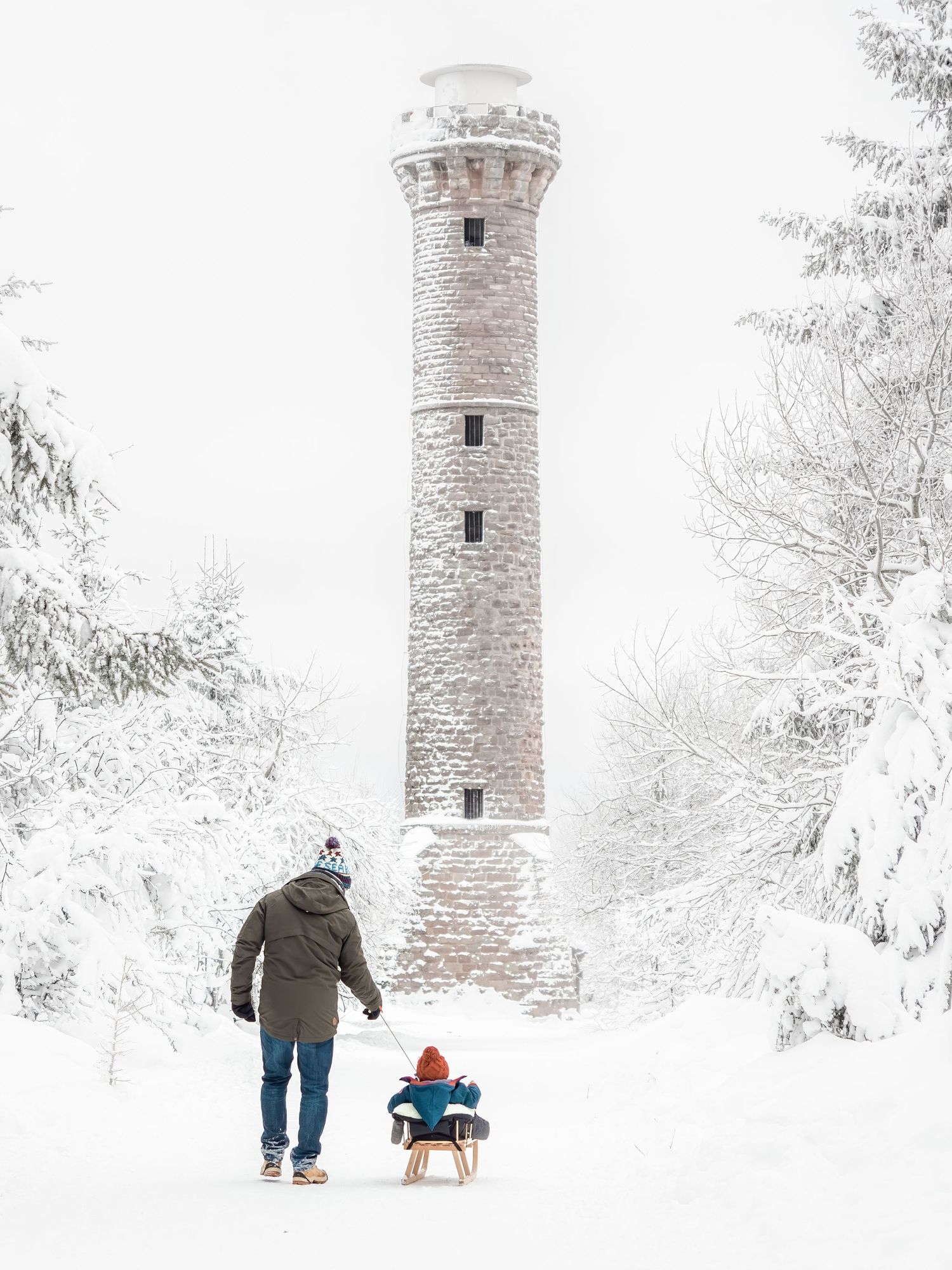 Ein Mann zieht einen Schlitten mit Kind, Ansicht von hinten, im Hintergrund ist ein Turm, die Landschaft ist schneebedekt