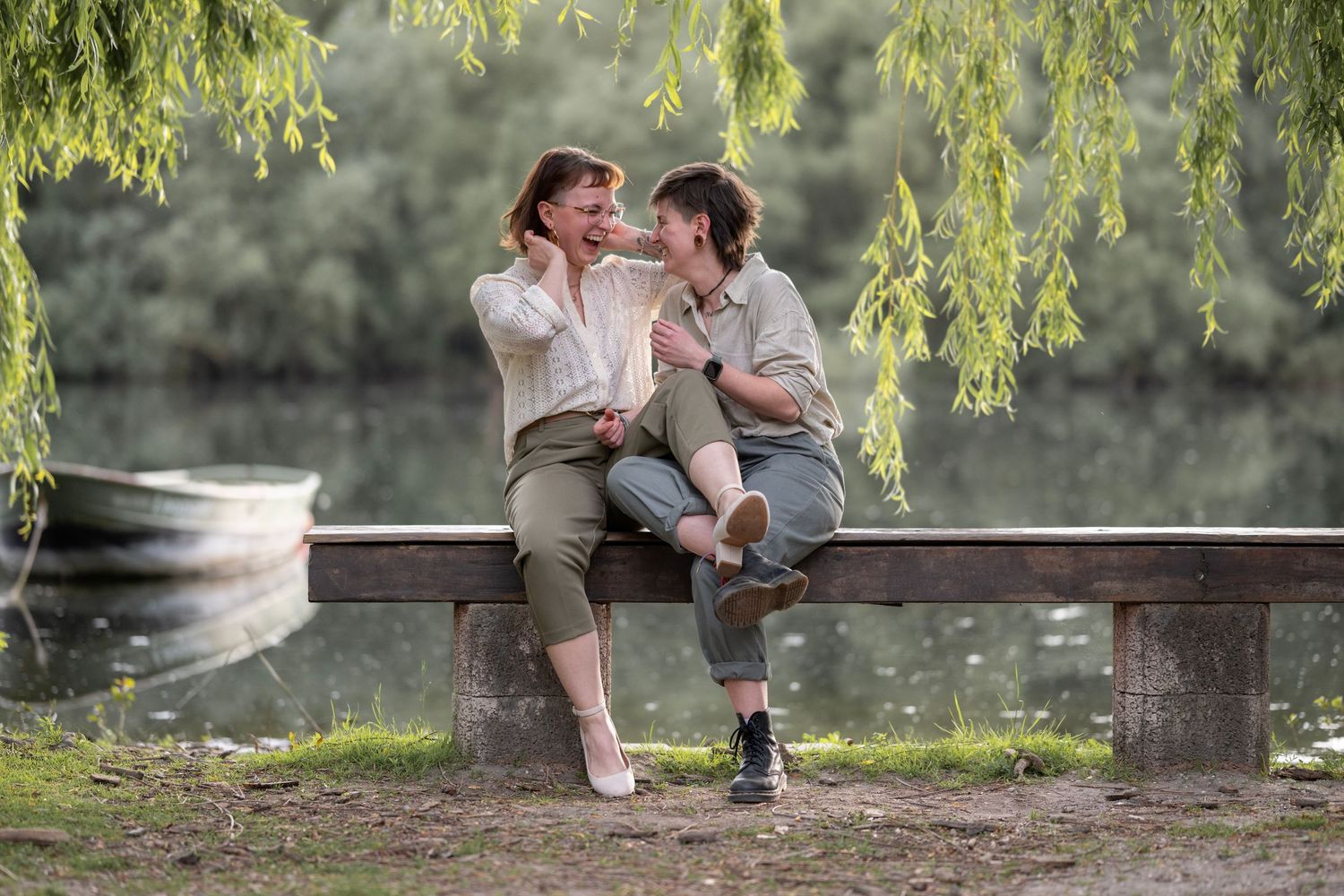 Foto von zwei Frauen die eng umschlungen auf einer Bank sitzen vor Wasser, lachen ausgelassen