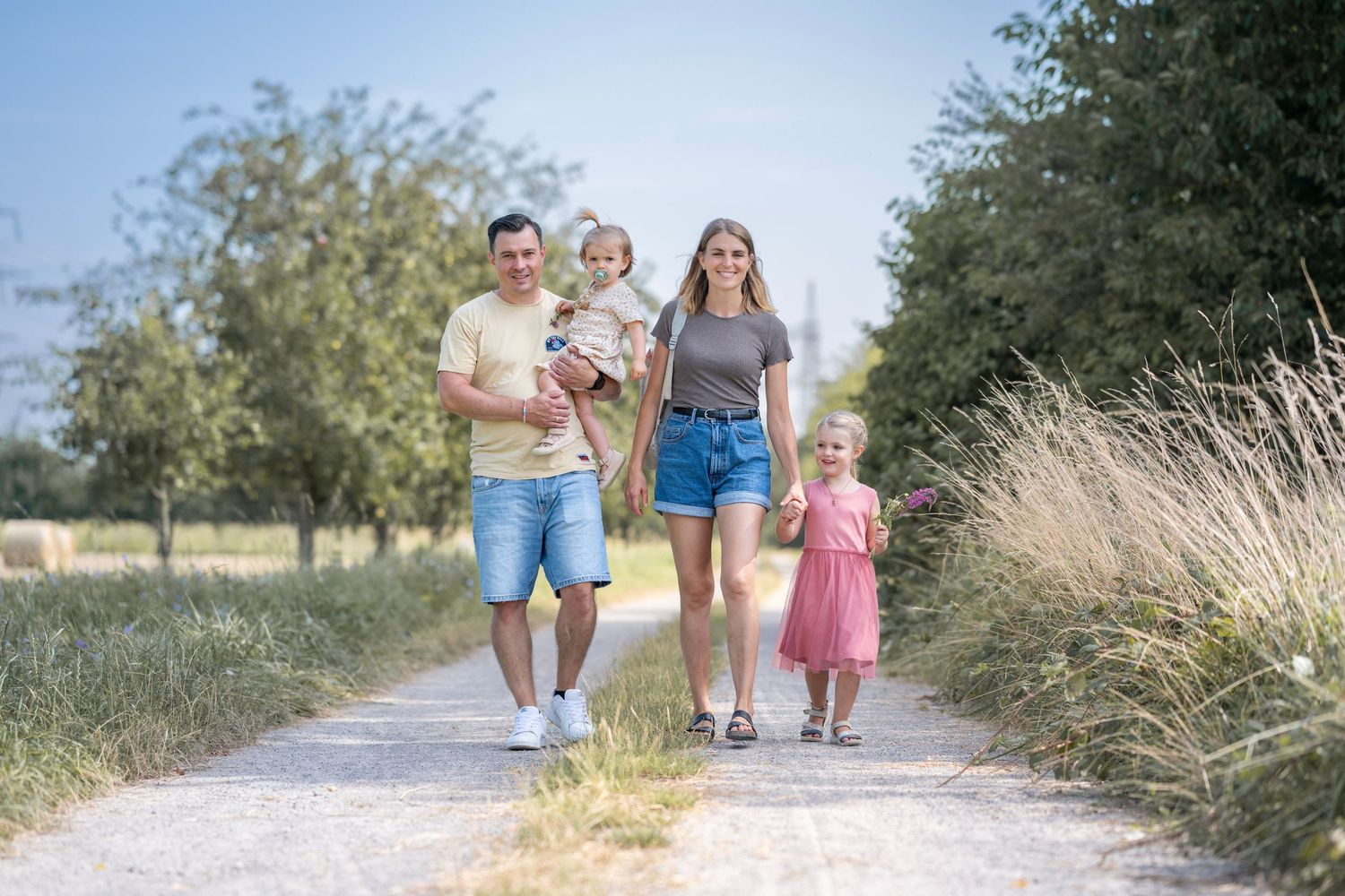 Eltern mit zwei Kindern laufen auf einem Feldweg. Der Vater trägt ein Kind, die Mutter führt eins an der Hand