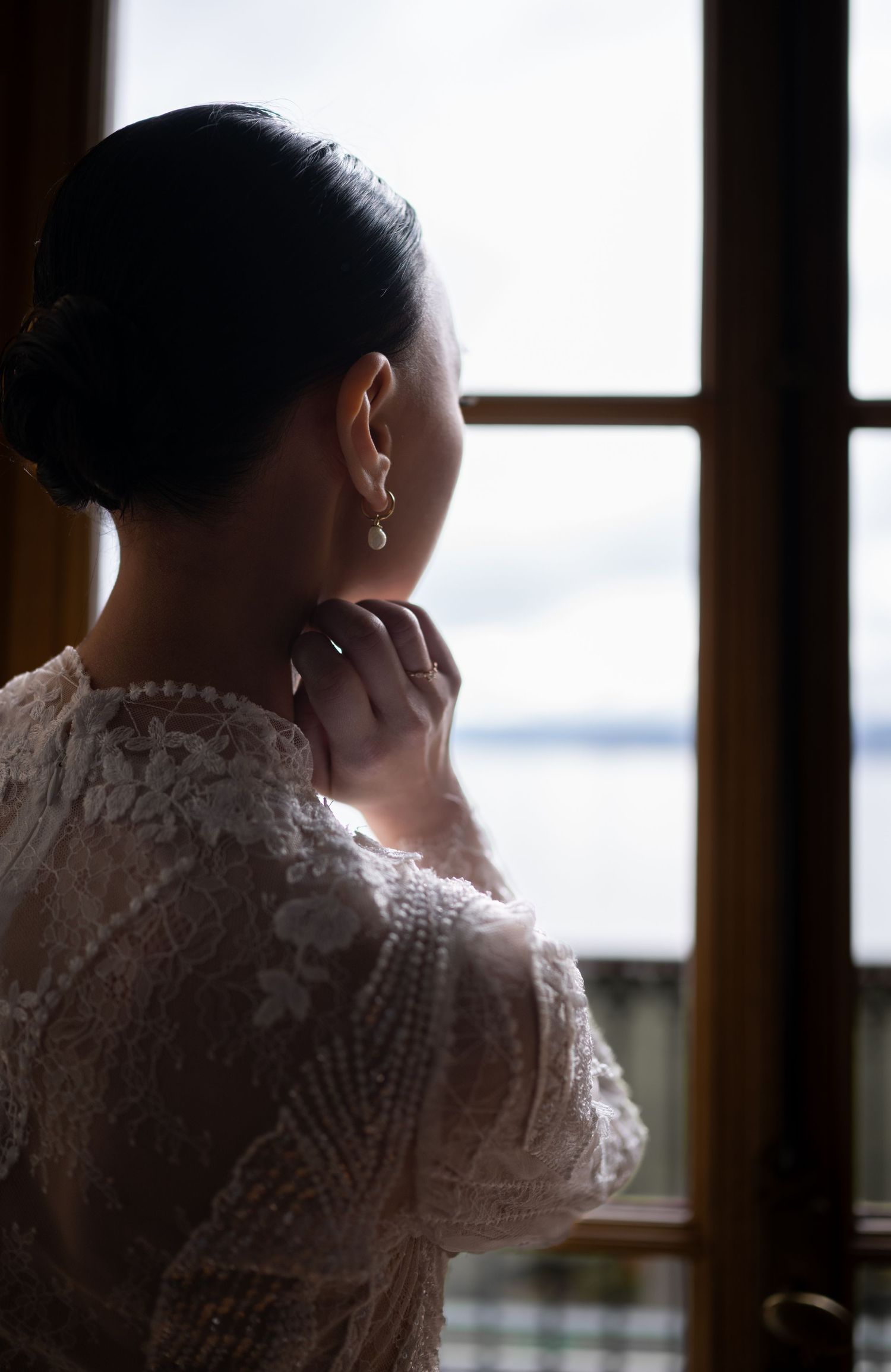 Eine Frau im Brautkleid schaut aus dem Fenster, fasst sich an den Hals, mit Perlen Ohrring