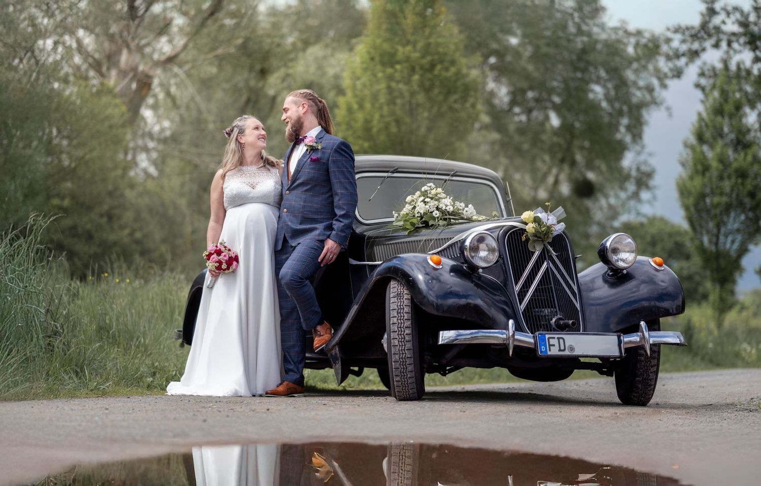 Foto vom Brautpaar angelehnt am Oldtimer, geschmückt mit Blumen