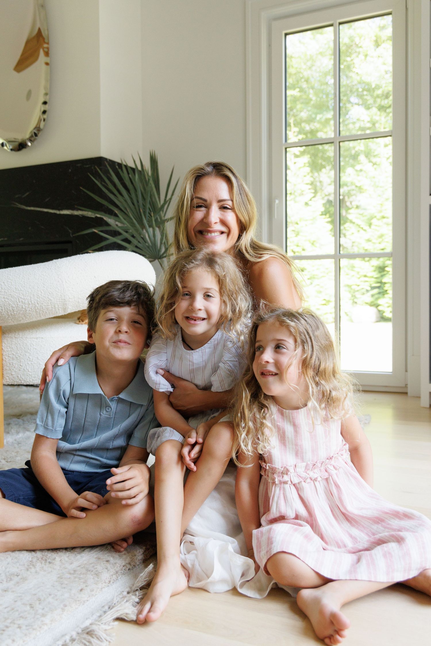 Siblings lounging together on white carpet in sunlit room with large windows.