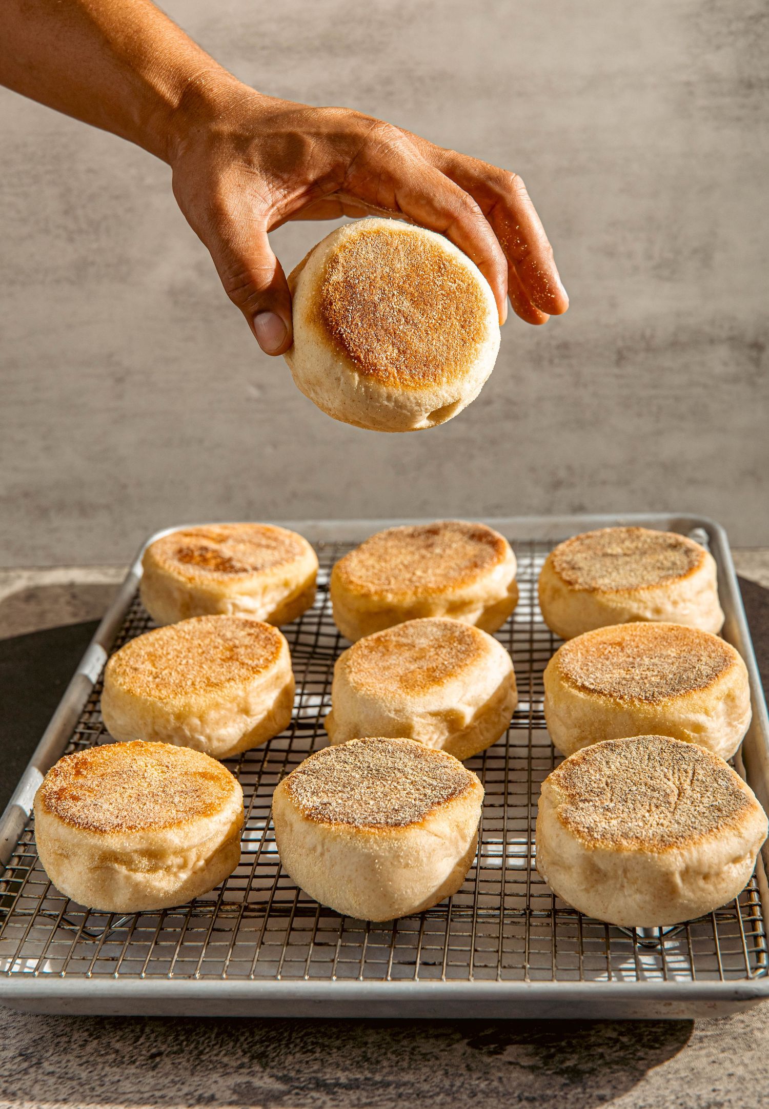 Homemade English muffins arranged on a cooling rack with one being picked up.