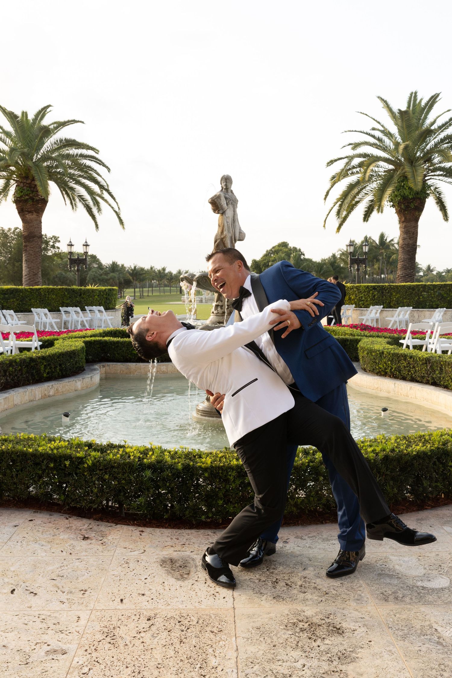 Two people in formal attire playfully pose by a fountain surrounded by palm trees and white chairs.
