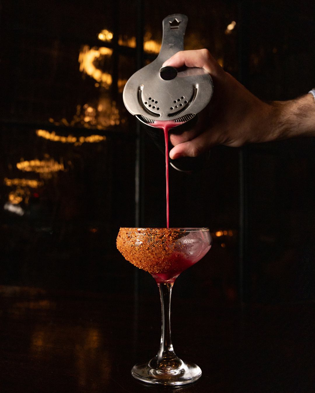 Craft cocktail being strained into a sugar-rimmed martini glass with dramatic dark background lighting.