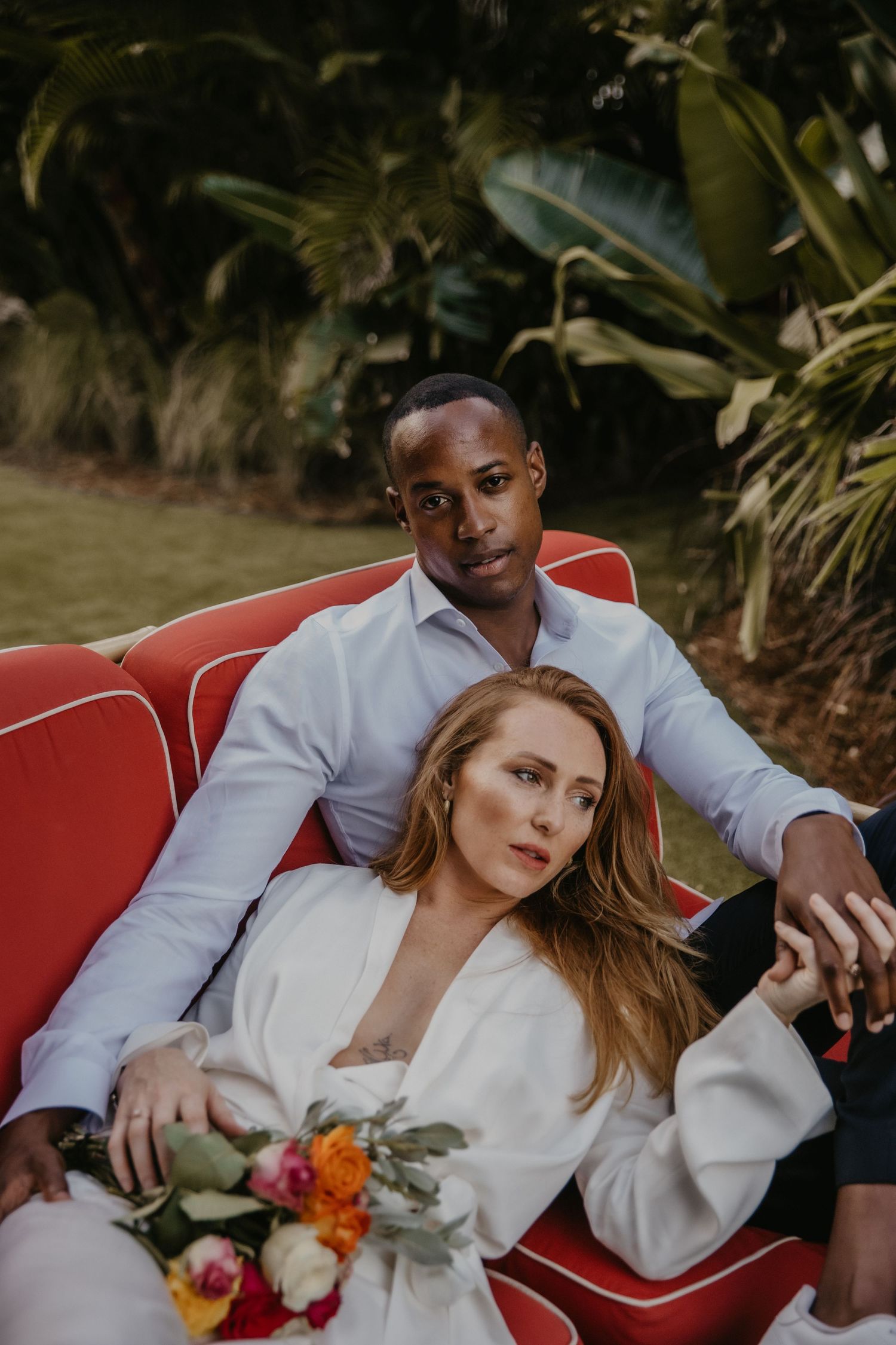 Romantic couple lounging on a red vintage couch in a garden setting.