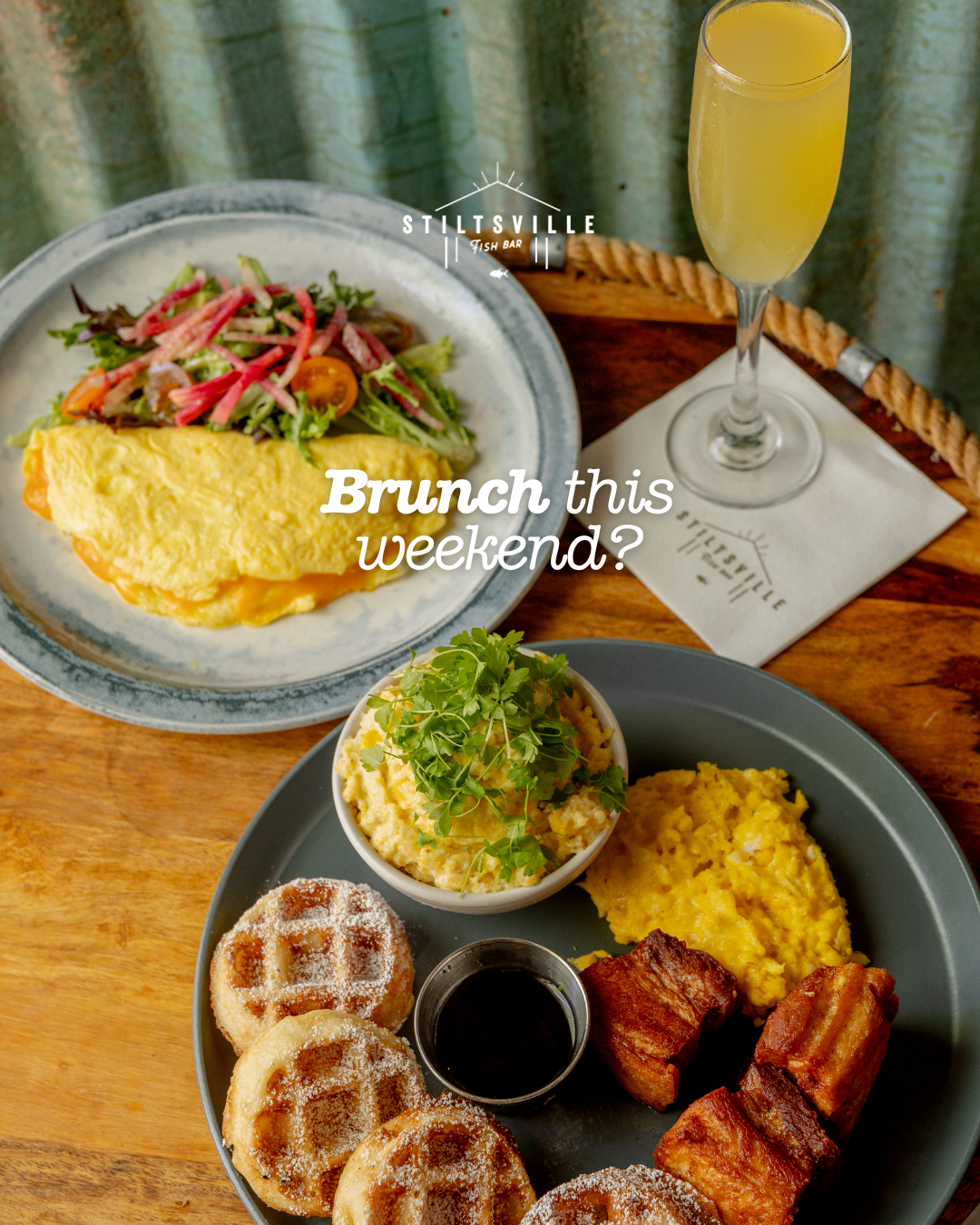 Brunch spread featuring fluffy omelettes, waffles, bacon and a mimosa on a wooden table.