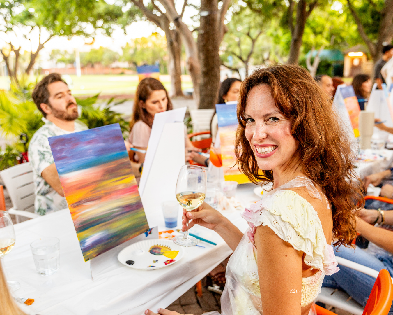 A group of people enjoying an outdoor dinner party with colorful blankets and wine glasses at sunset.