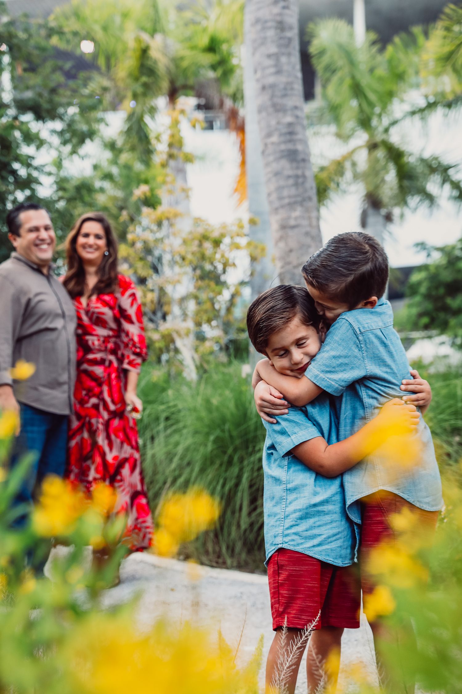 Family members sharing warm embraces in a garden with yellow flowers and palm trees.